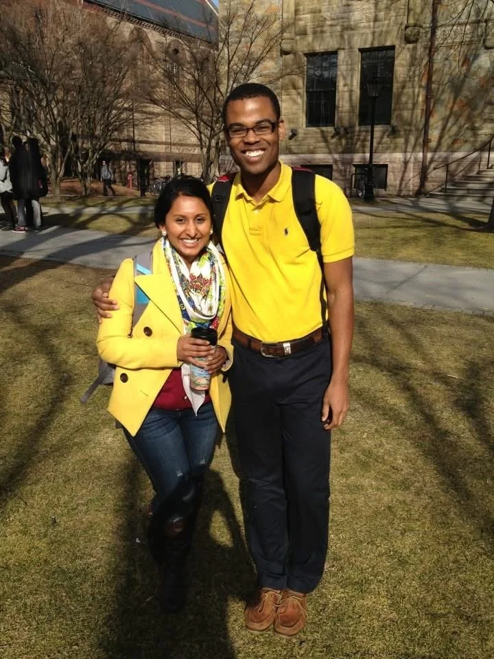 Two people, a woman and a man, standing on grass in a park or campus with leafless trees and a stone building in the background. The woman is wearing a yellow jacket, patterned scarf, and holding a paper coffee cup. The man is wearing a yellow polo s