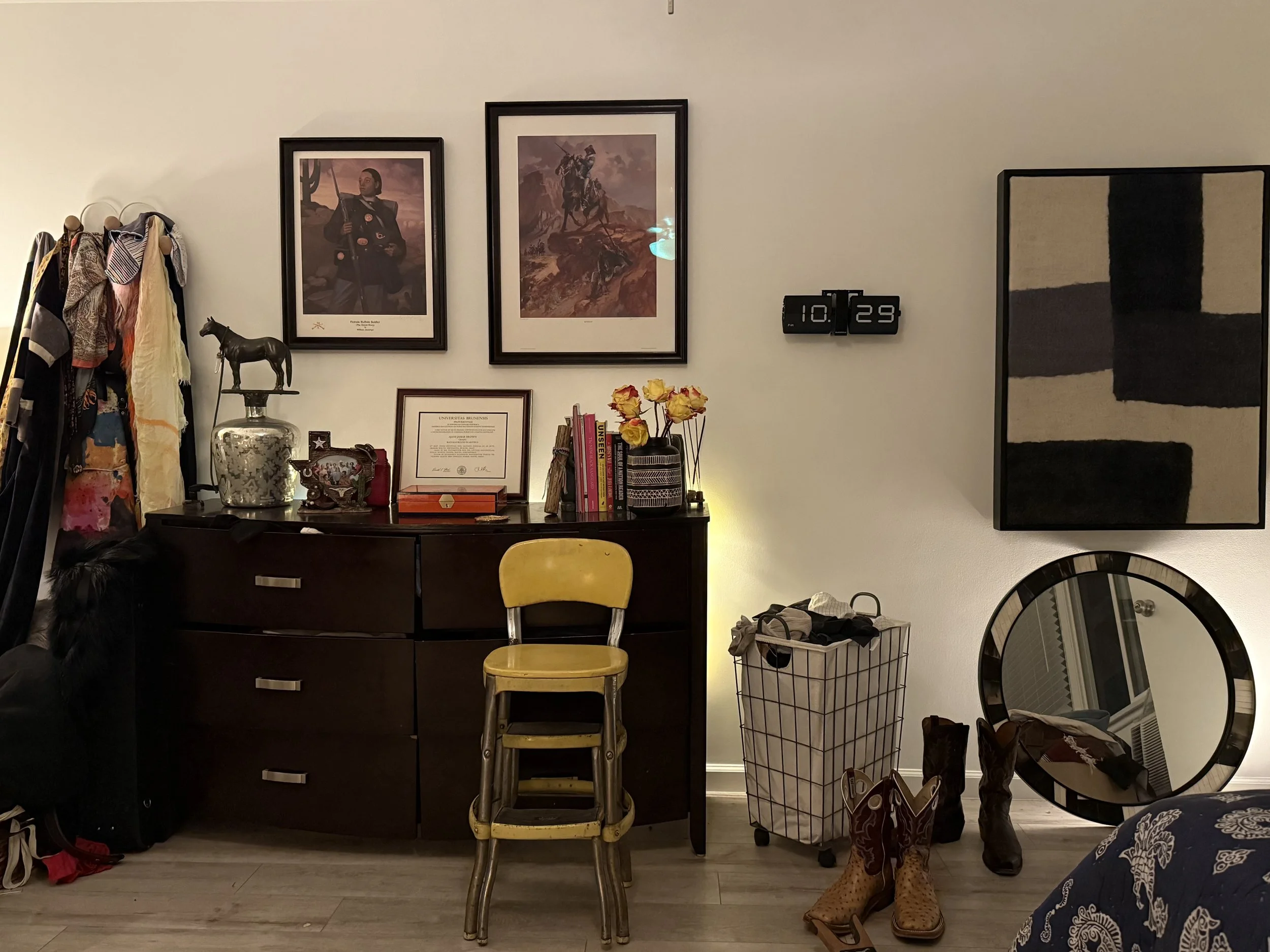 Living room corner with a dark wooden sideboard decorated with framed photographs and certificates, books, flowers, and decorative items. A yellow chair is in front of the sideboard. To the right, there are shoes, boots, and a large round mirror lean