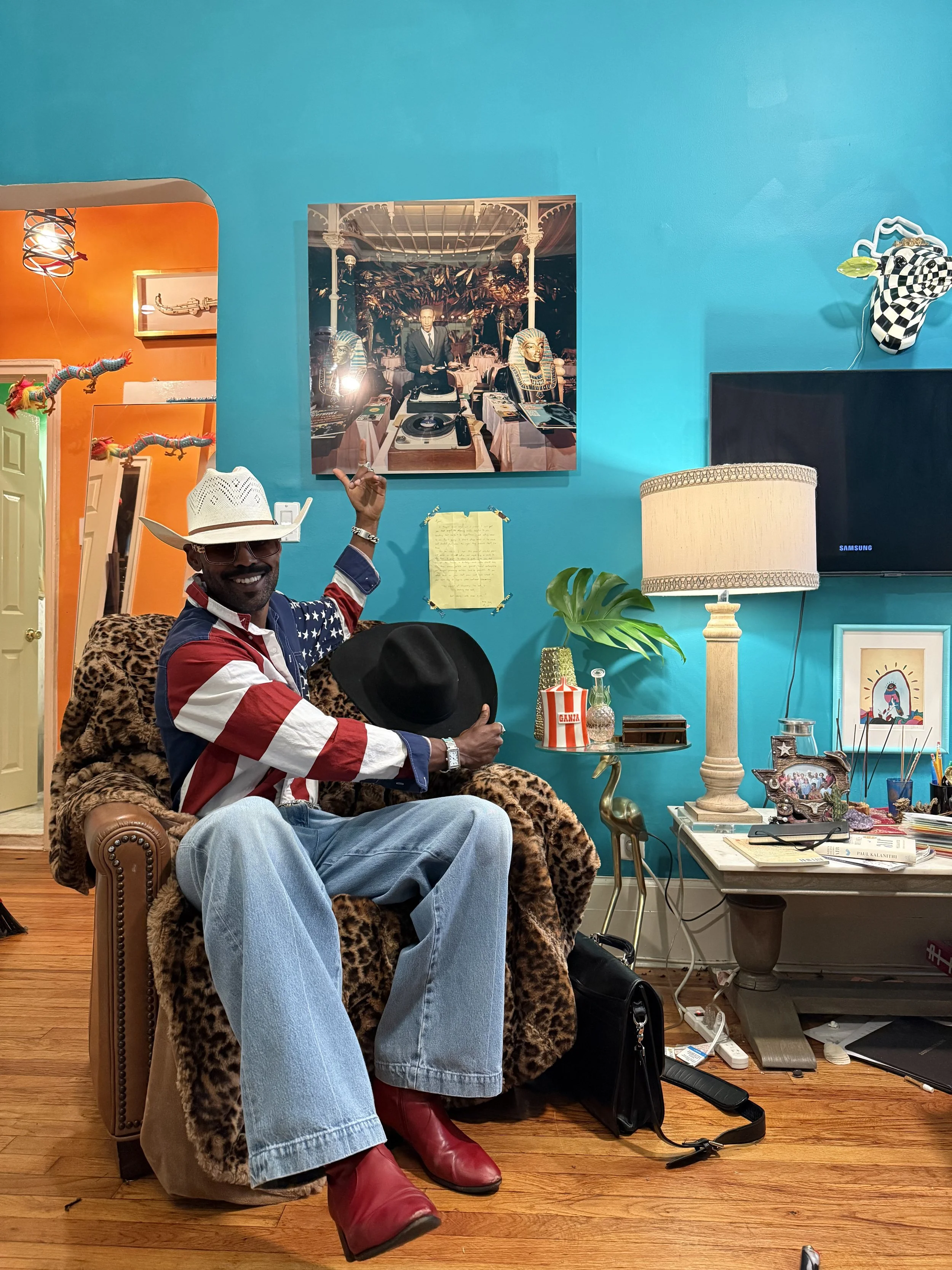 A man sitting on a leopard-print armchair in a colorful, eclectic room. He is smiling, wearing sunglasses, a white cowboy hat, a patriotic American flag shirt, light blue jeans, and red boots. The man is holding a black hat and pointing to a picture