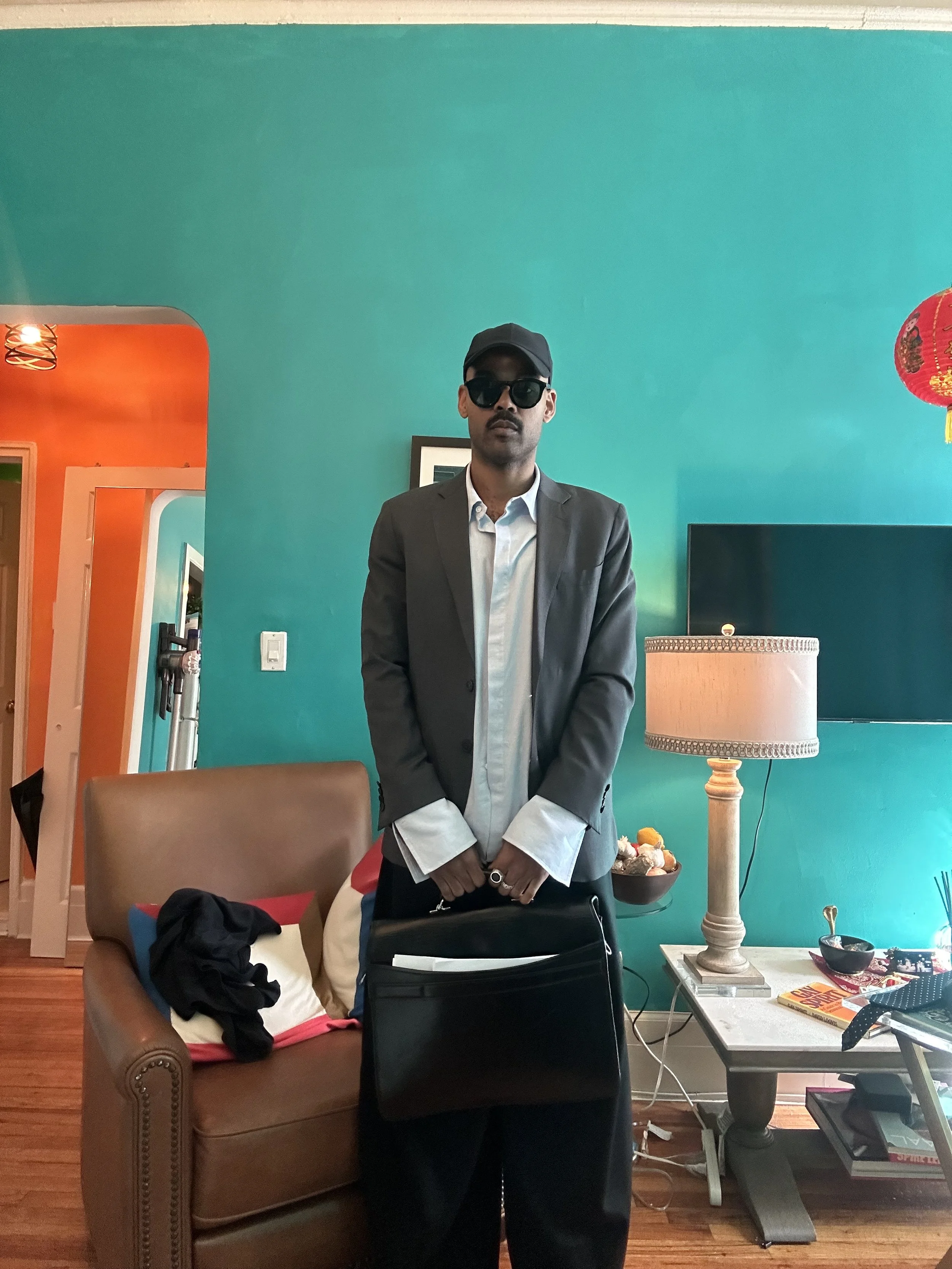 A young man dressed in a blazer, white shirt, sunglasses, and cap standing in a colorful living room with a teal wall, a brown leather chair, a side table with a lamp, and various objects.