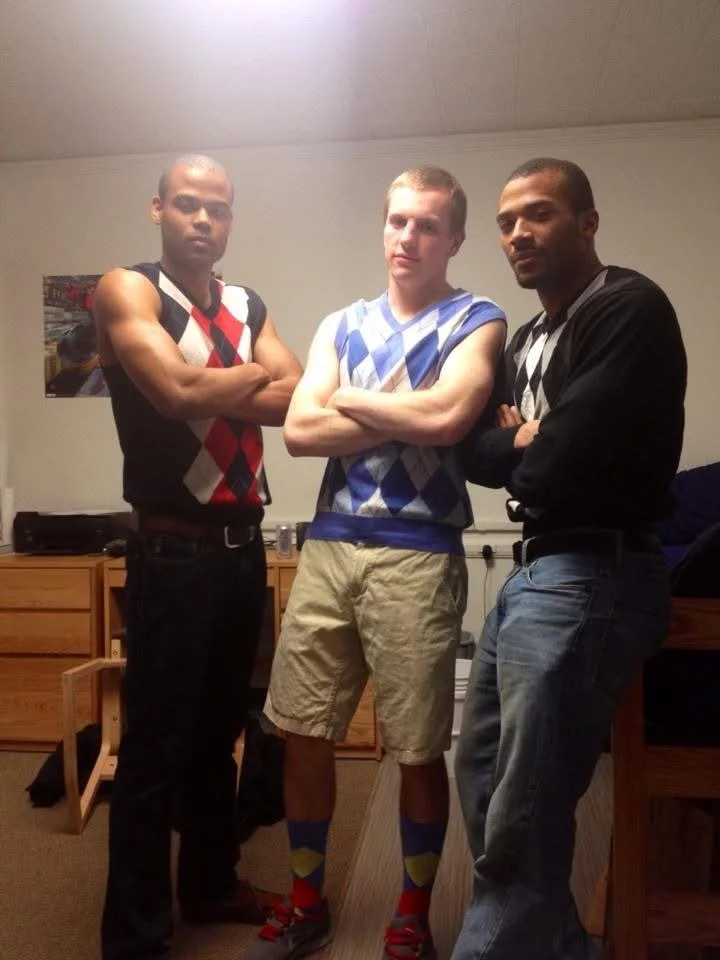 Three young men standing with crossed arms; two wearing sleeveless argyle sweater vests and the middle man wearing a team sports jersey; they are indoors in a room with wooden furniture.