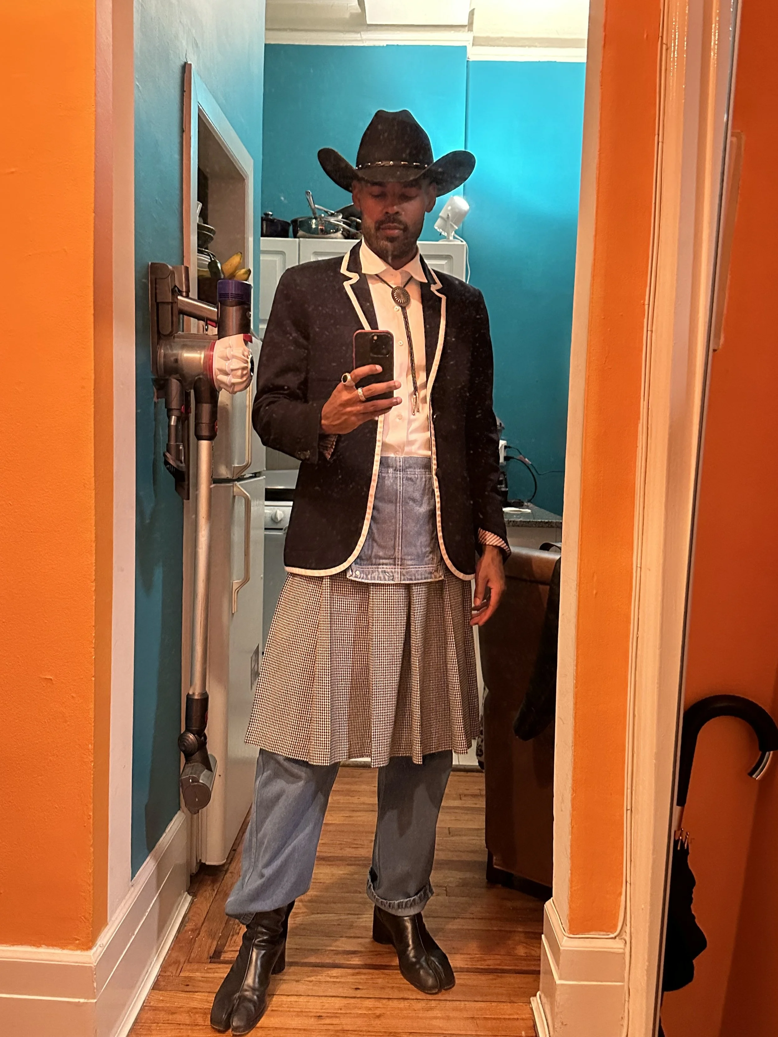 Man taking a mirror selfie dressed in a cowboy hat, blazer, dress shirt, skirt, jeans, and cowboy boots.