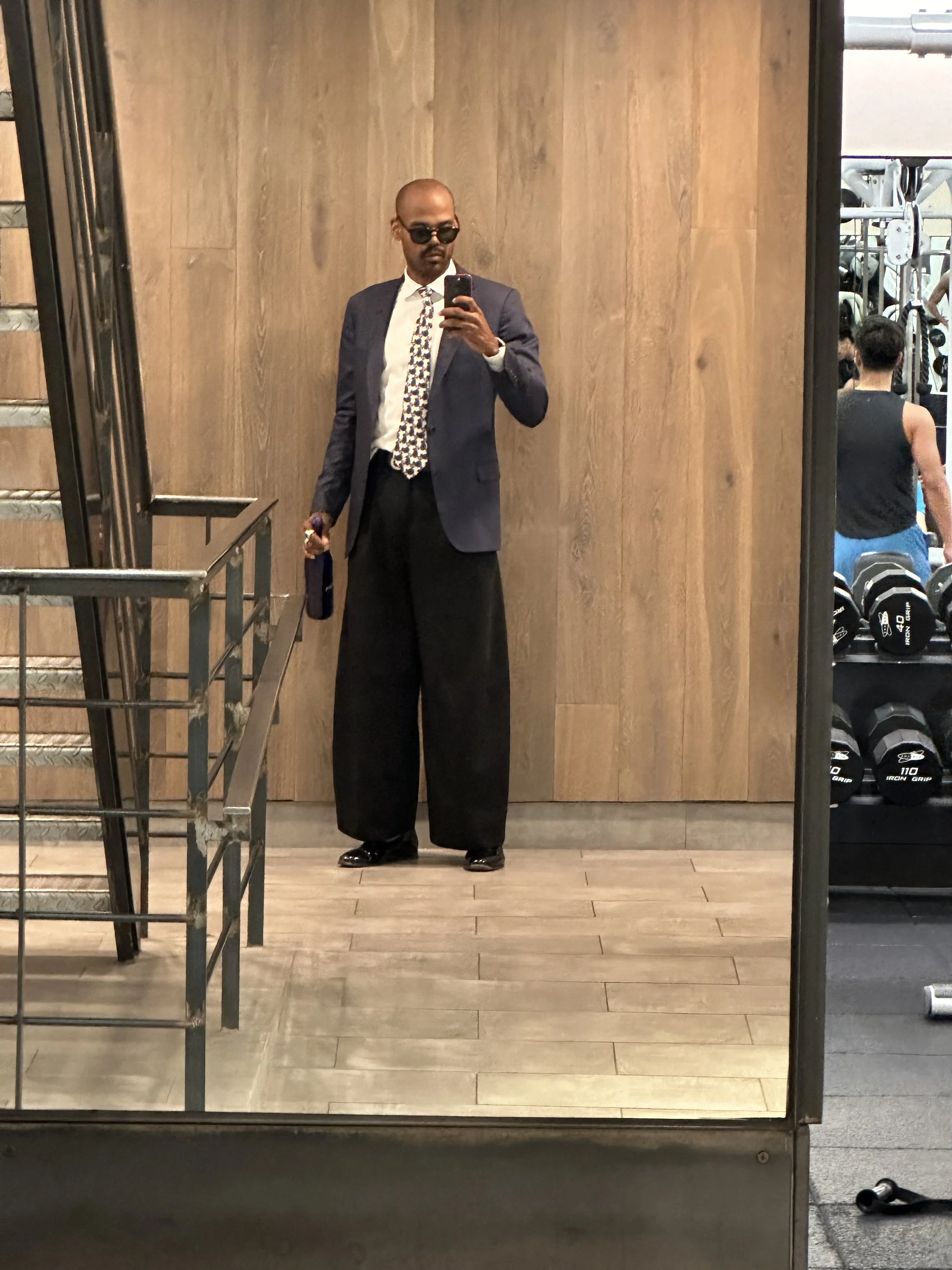 A man taking a mirror selfie in a gym locker room. He is dressed in a navy blue blazer, white shirt, patterned tie, large black pants, and sunglasses. He is holding a water bottle and appears to be in a workout facility.