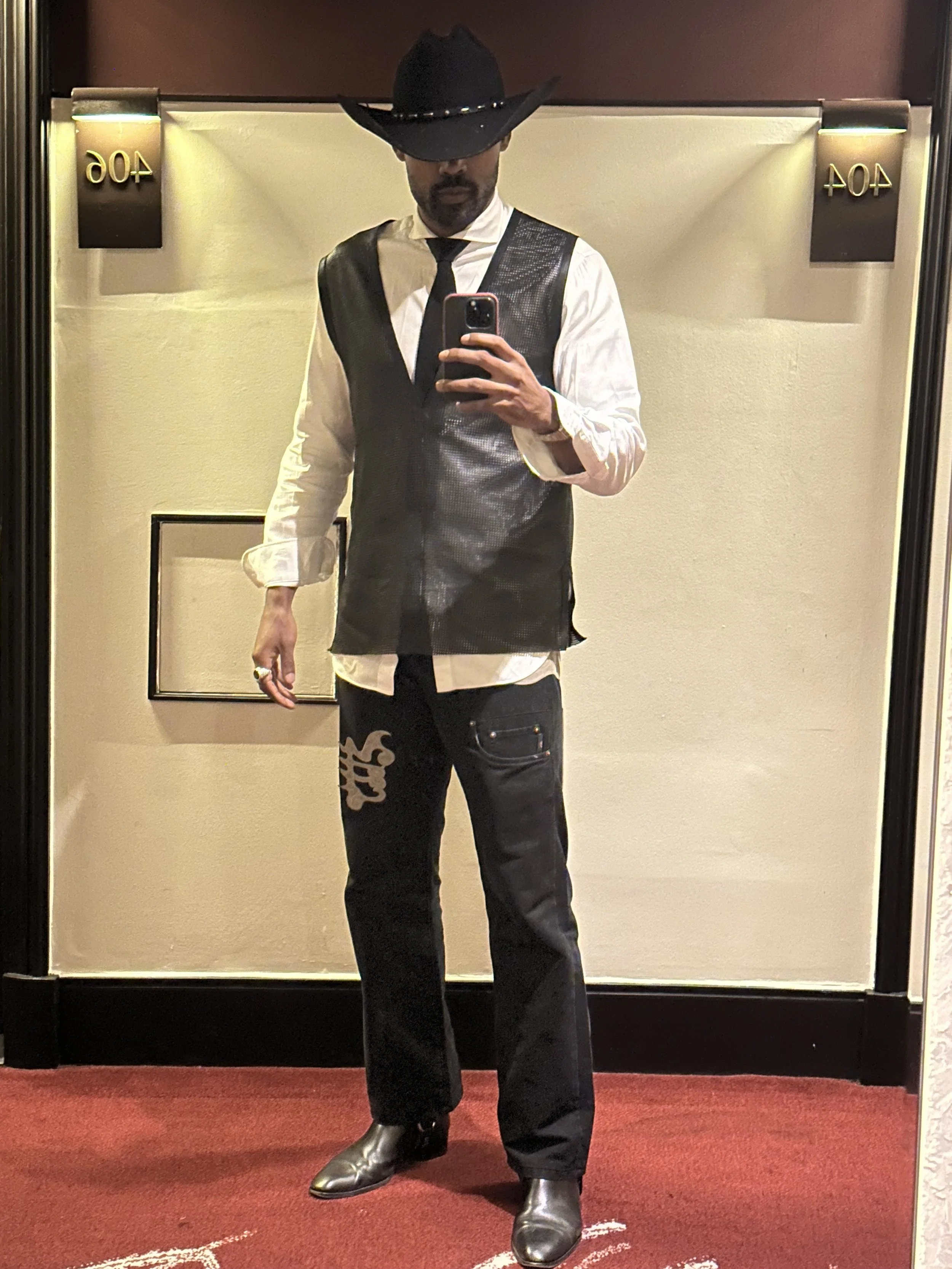A man taking a mirror selfie in a hallway. He's wearing a black cowboy hat, a white shirt, a black vest, a black tie, black jeans with white text on the leg, and black boots.