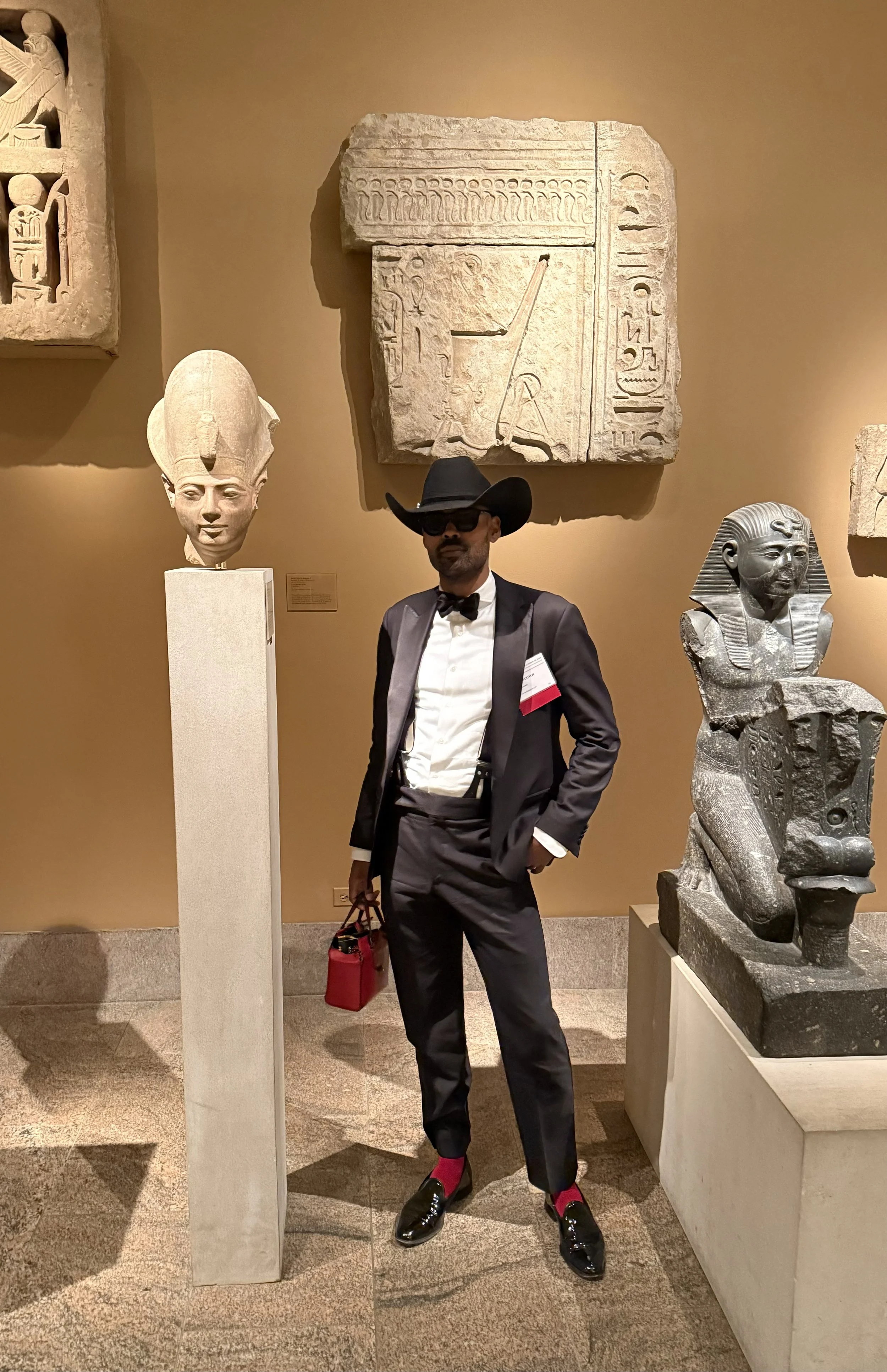 A man dressed in a tuxedo with a bow tie, black hat, sunglasses, red socks, and black shoes stands in an art museum next to ancient Egyptian sculptures and carvings displayed on a beige wall.
