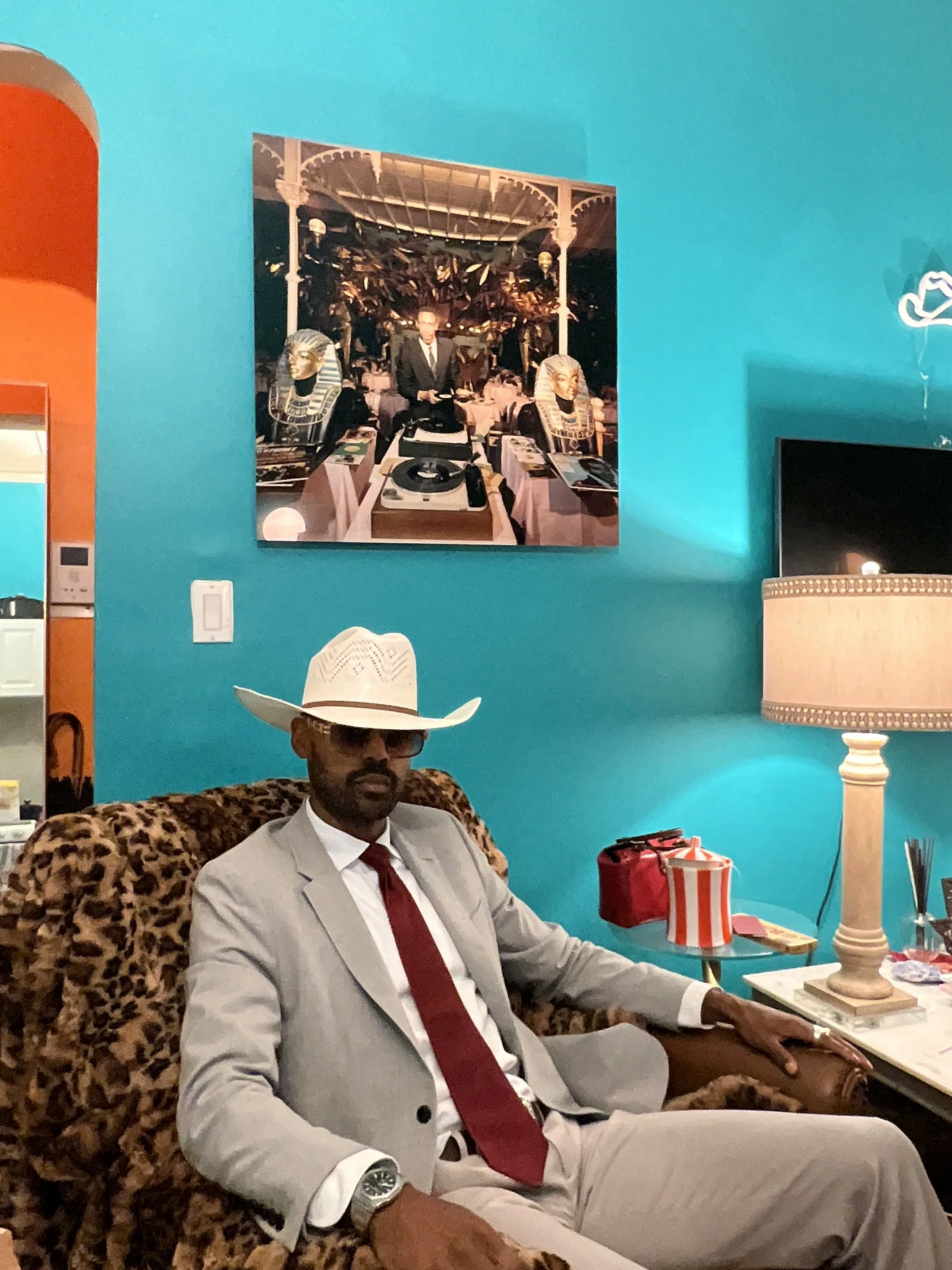 A man dressed in a light gray suit, white shirt, red tie, white cowboy hat, sunglasses, and watch, sitting on a leopard-print armchair in a room with turquoise walls, a beige lamp, and a wall art picture of a man in a black suit at a buffet table. Th