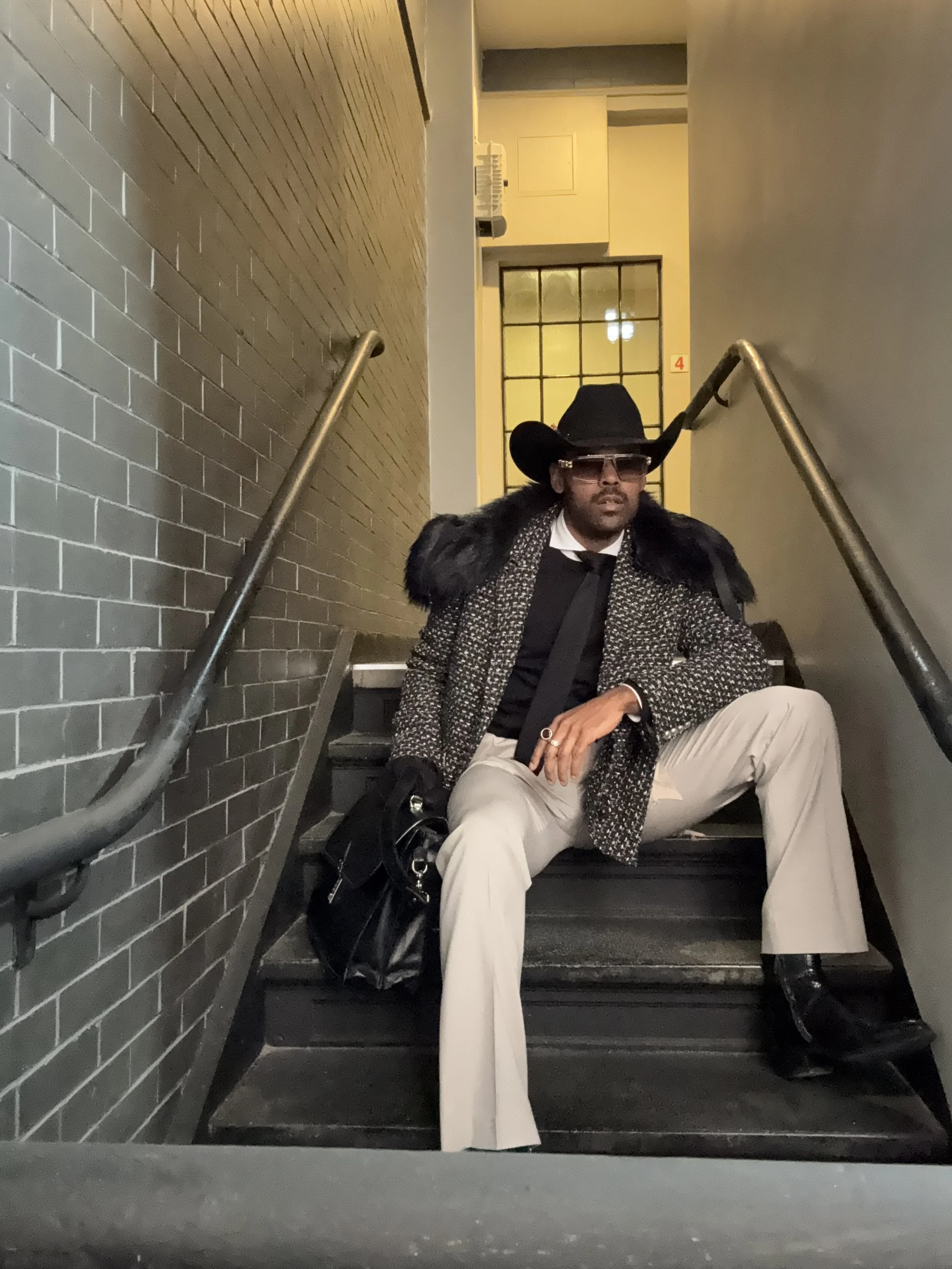 A man sitting on stairs, dressed in a fashionable outfit with a black wide-brimmed hat, sunglasses, a patterned coat with fur collar, black shoes, beige pants, and holding a black bag.