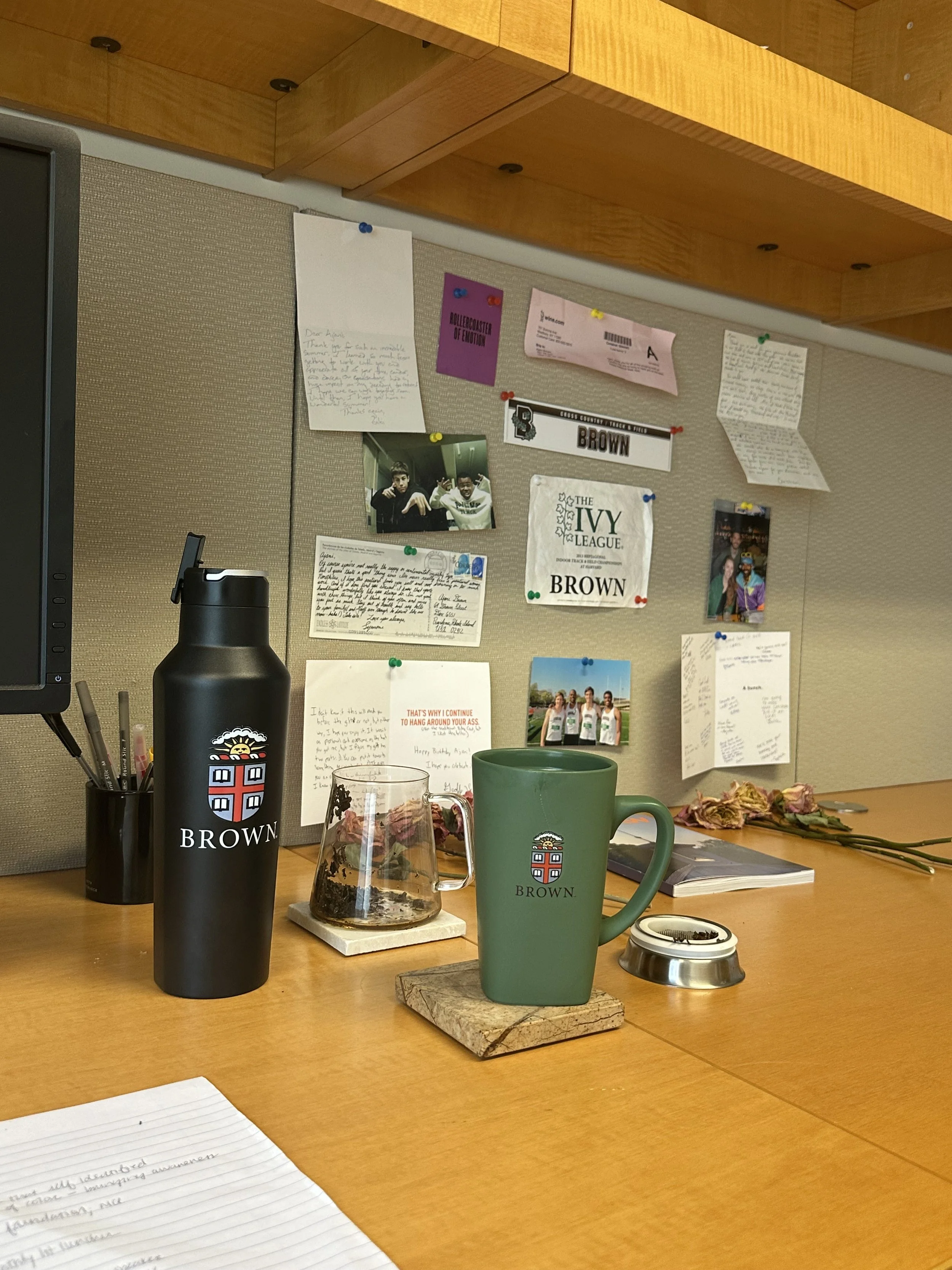 Office desk with a green mug, a black water bottle, a glass with dried flowers, and a notebook. The background features a corkboard with various notes, photos, and memorabilia pinned up.