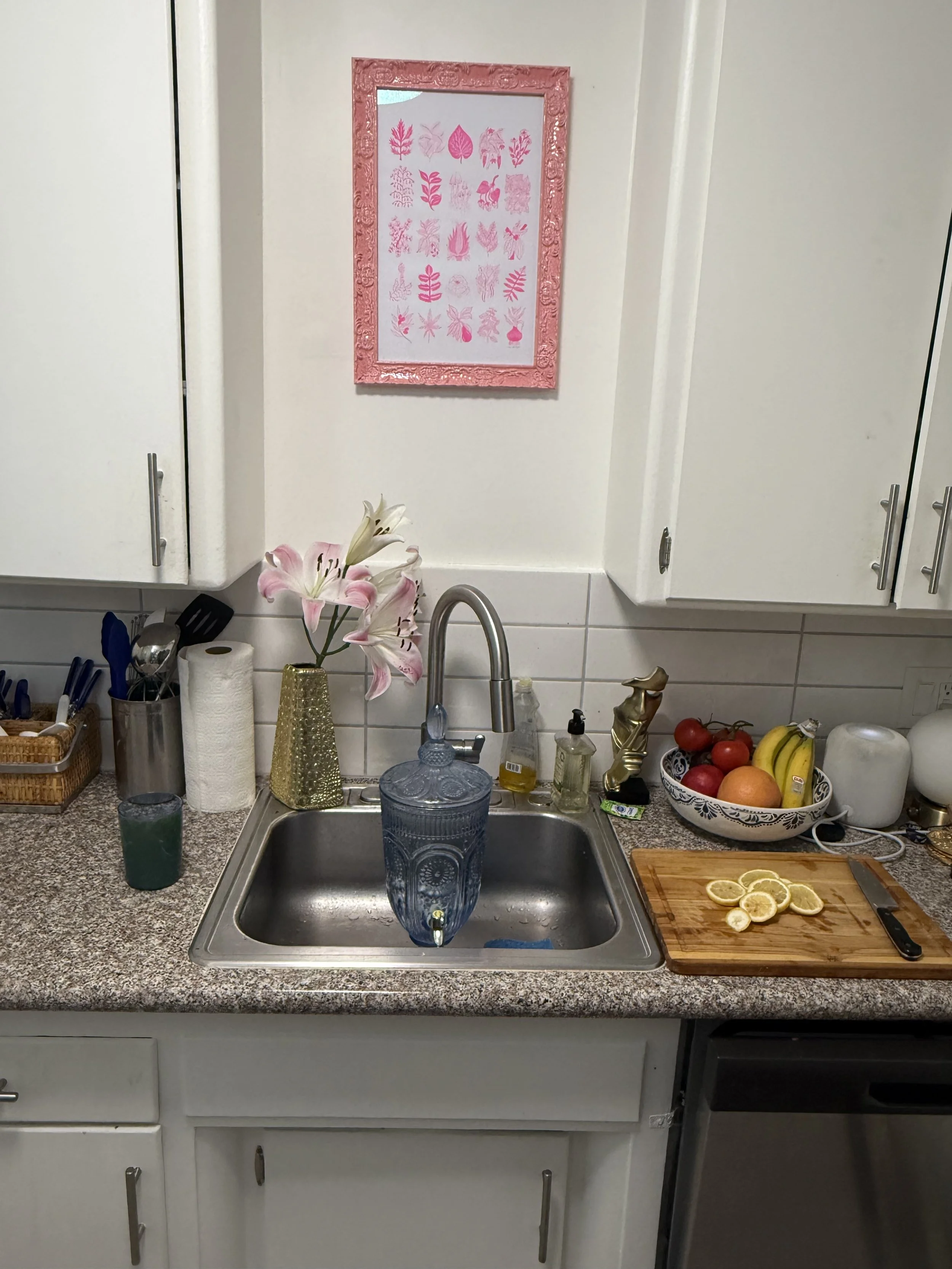 Kitchen sink with pink lilies in a gold vase, a cutting board with lemon slices, and a bowl of fruit including bananas, apples, and oranges. White cabinets and a pink framed art piece on the wall.