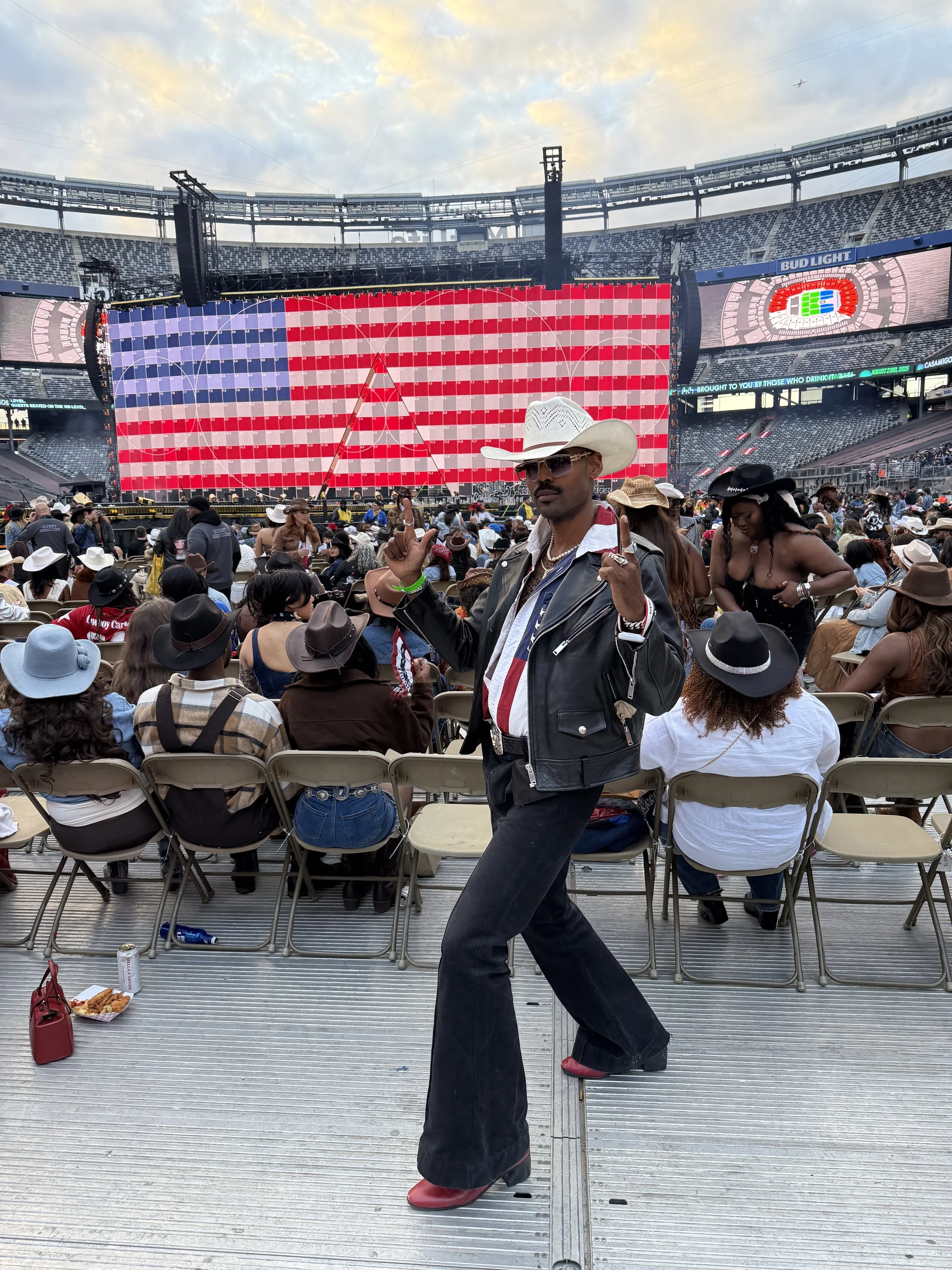 A man dressed in a cowboy hat, sunglasses, leather jacket, and bell-bottom jeans posing with a peace sign at a concert or festival in a stadium with a large LED display and crowd in the background.
