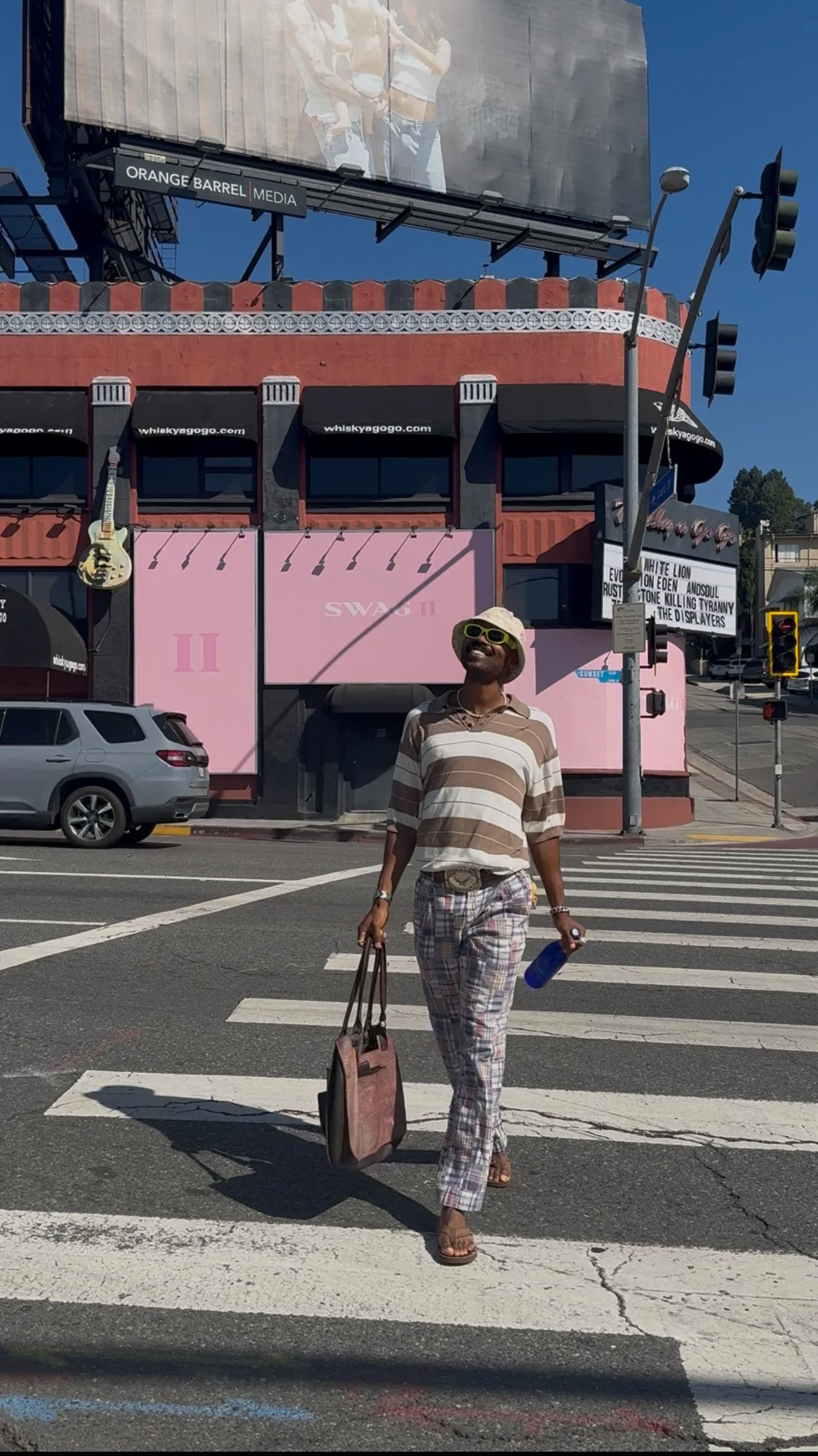 A woman crossing the street at a crosswalk, smiling and wearing sunglasses, a striped beige and white shirt, plaid pants, and sandals, carrying a tote bag in one hand and a water bottle in the other, with a pink building, billboard, and traffic light