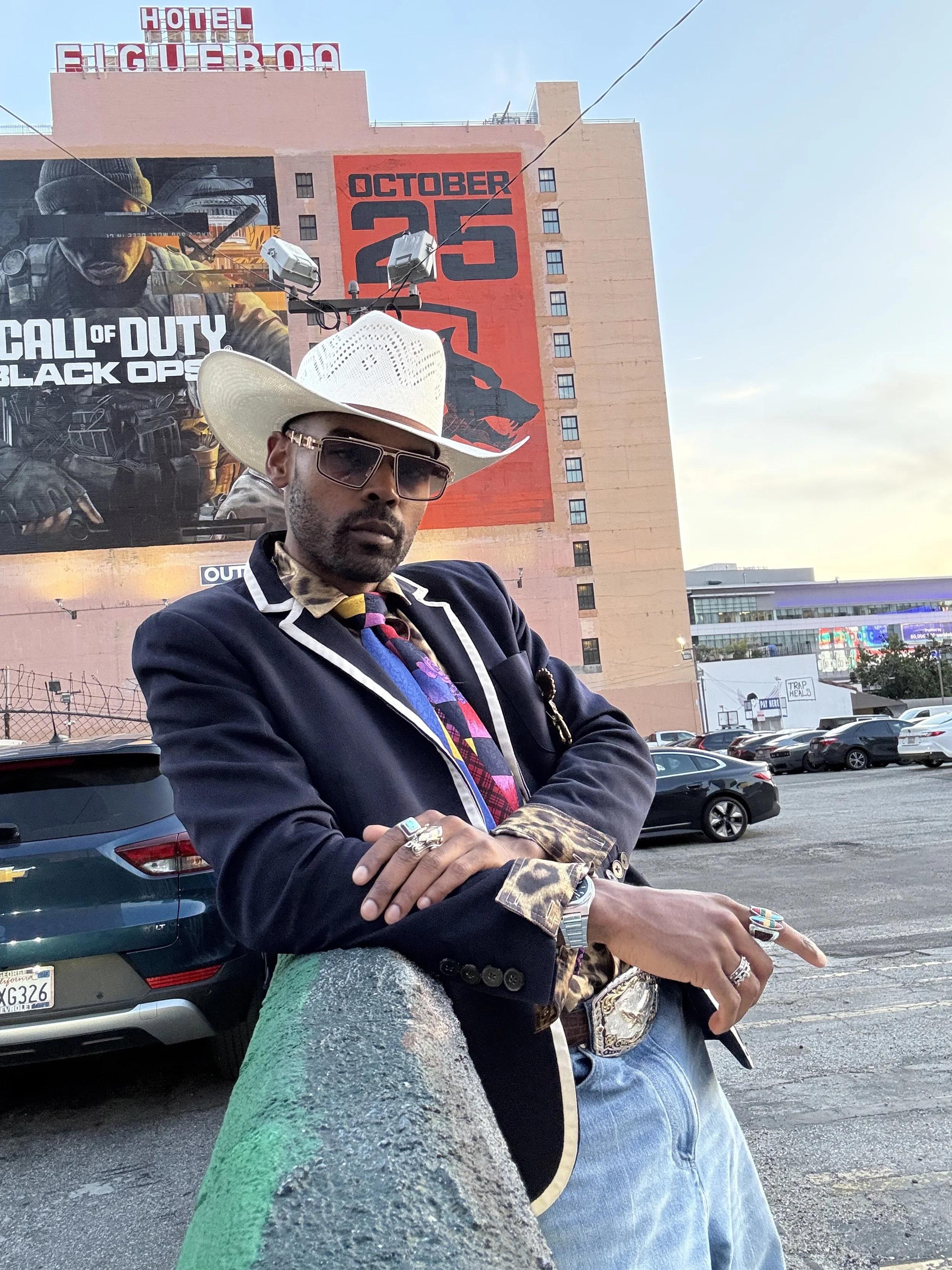 A man wearing a white cowboy hat, sunglasses, a dark blazer with leopard print details, and a colorful tie, leaning against a painted parking lot curb with a hotel and billboard in the background.