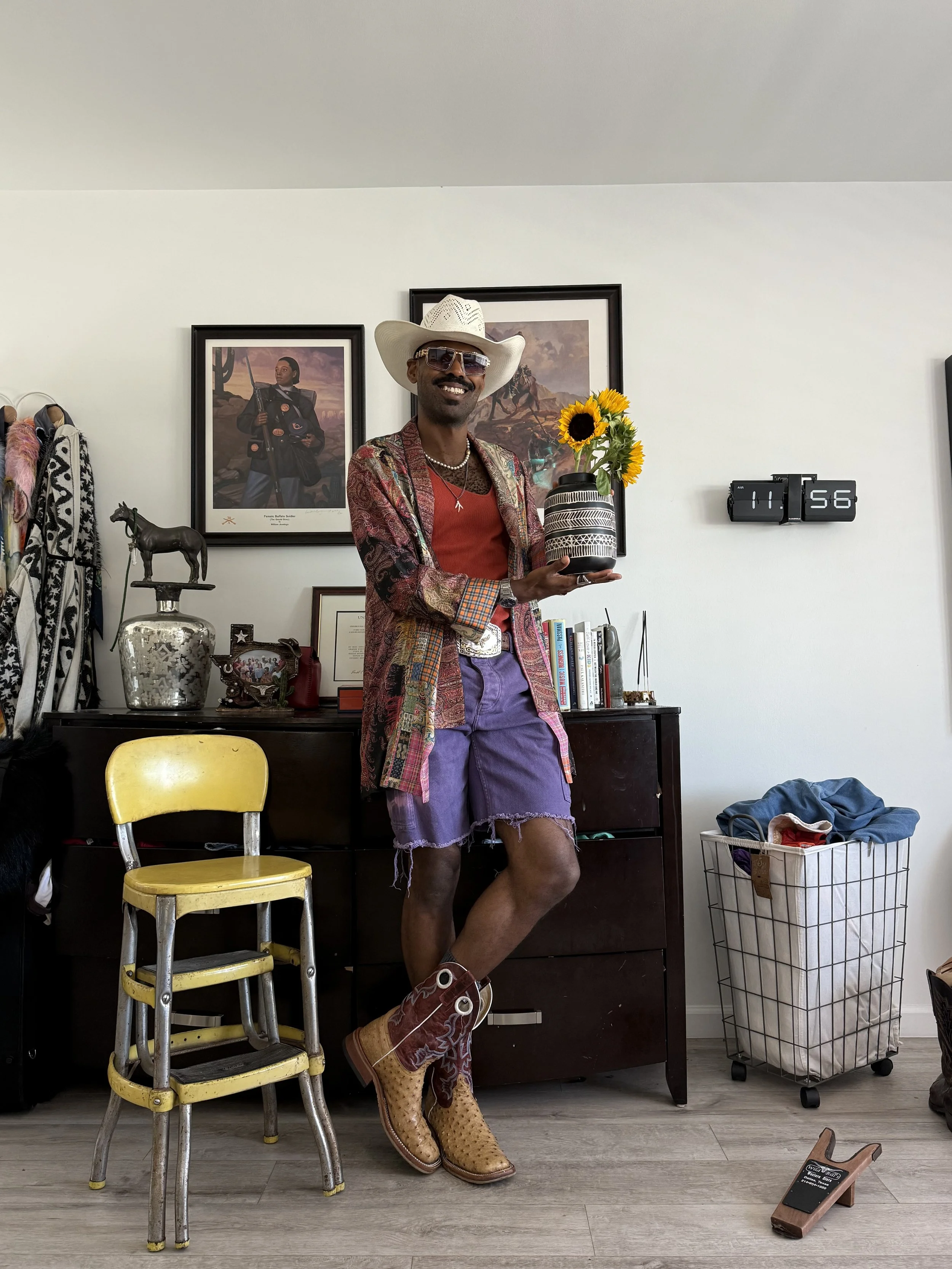 A man dressed in eclectic, colorful fashion standing in a room, holding a vase with sunflowers, wearing a cowboy hat, sunglasses, a patterned blazer, purple shorts, and cowboy boots, smiling