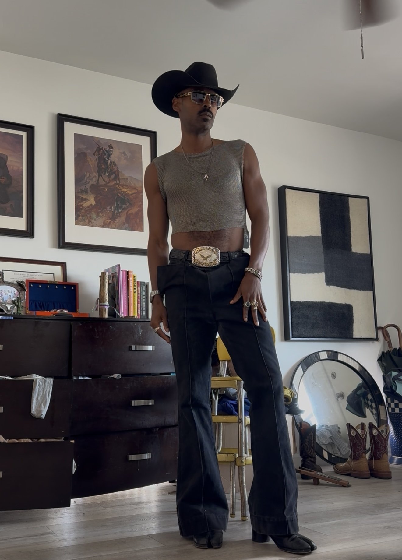 Person standing indoors wearing a wide-brimmed black hat, sunglasses, sleeveless cropped shirt, dark jeans with a large belt, and cowboy boots, with art and furniture in the background.