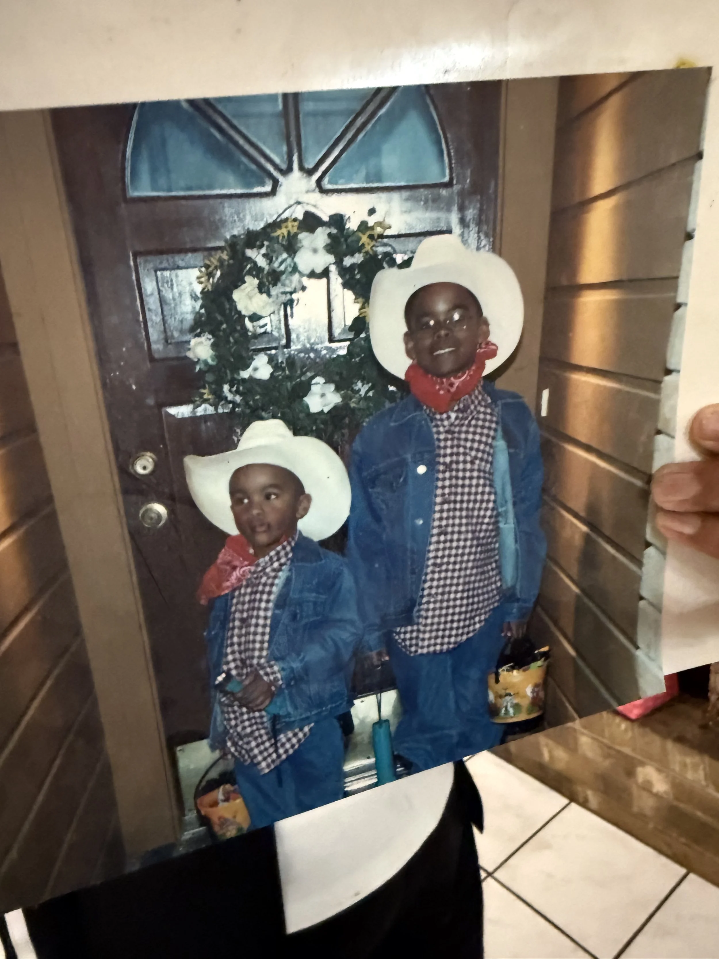 A photo of two young boys wearing cowboy hats, denim jackets, and checkered shirts, standing in front of a decorative wreath on a wooden door.