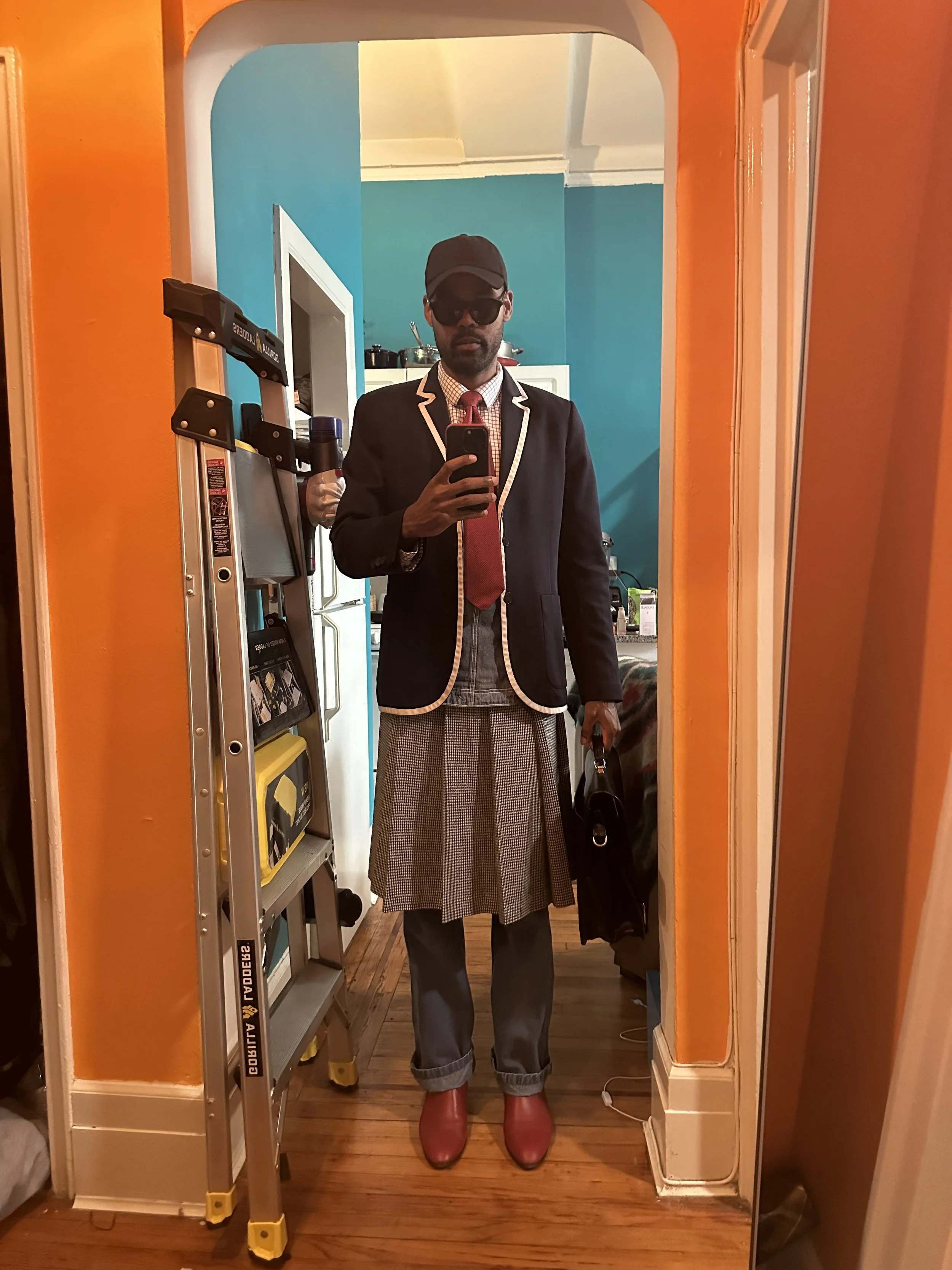 Person taking a mirror selfie in a brightly colored room, dressed in a stylish outfit with a blazer, tie, pleated kilt, cropped pants, red shoes, sunglasses, cap, and holding a bag.