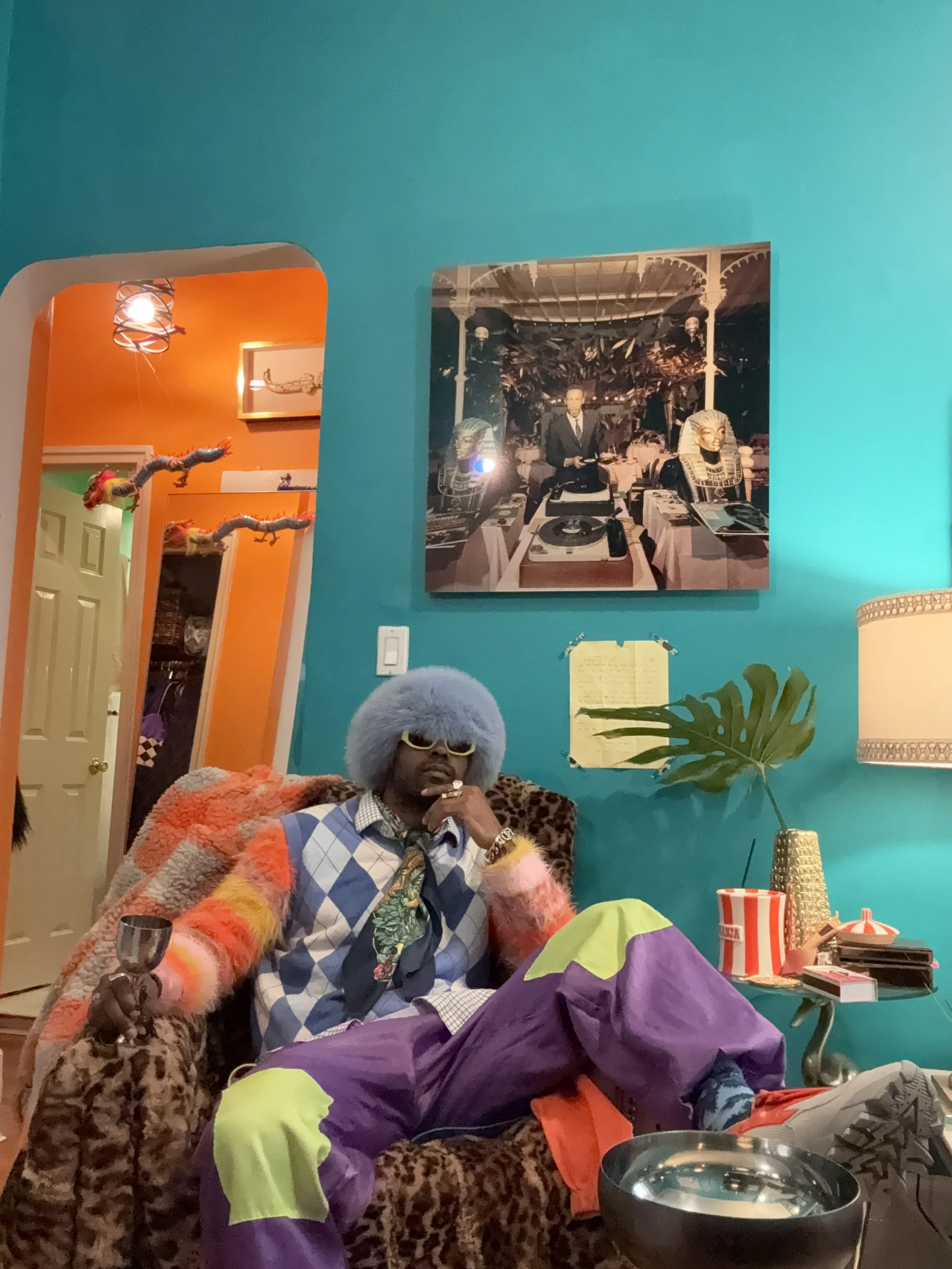 A person wearing a blue furry hat, sunglasses, and a colorful outfit sitting in a decorated room with teal walls, leopard print chairs, and vibrant decor.