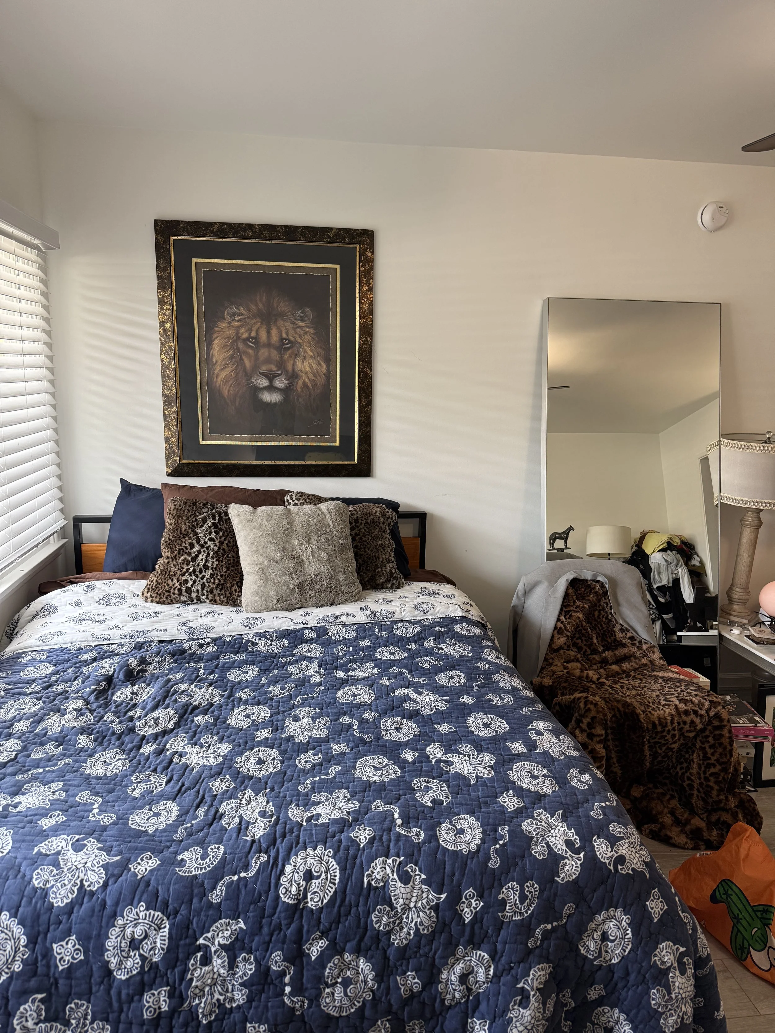 Bedroom with bed featuring a blue and white patterned quilt, and assorted pillows including animal print and fur. Above the bed, a framed lion portrait hangs on the wall. To the right, a large mirror leans against the wall, reflecting part of the roo