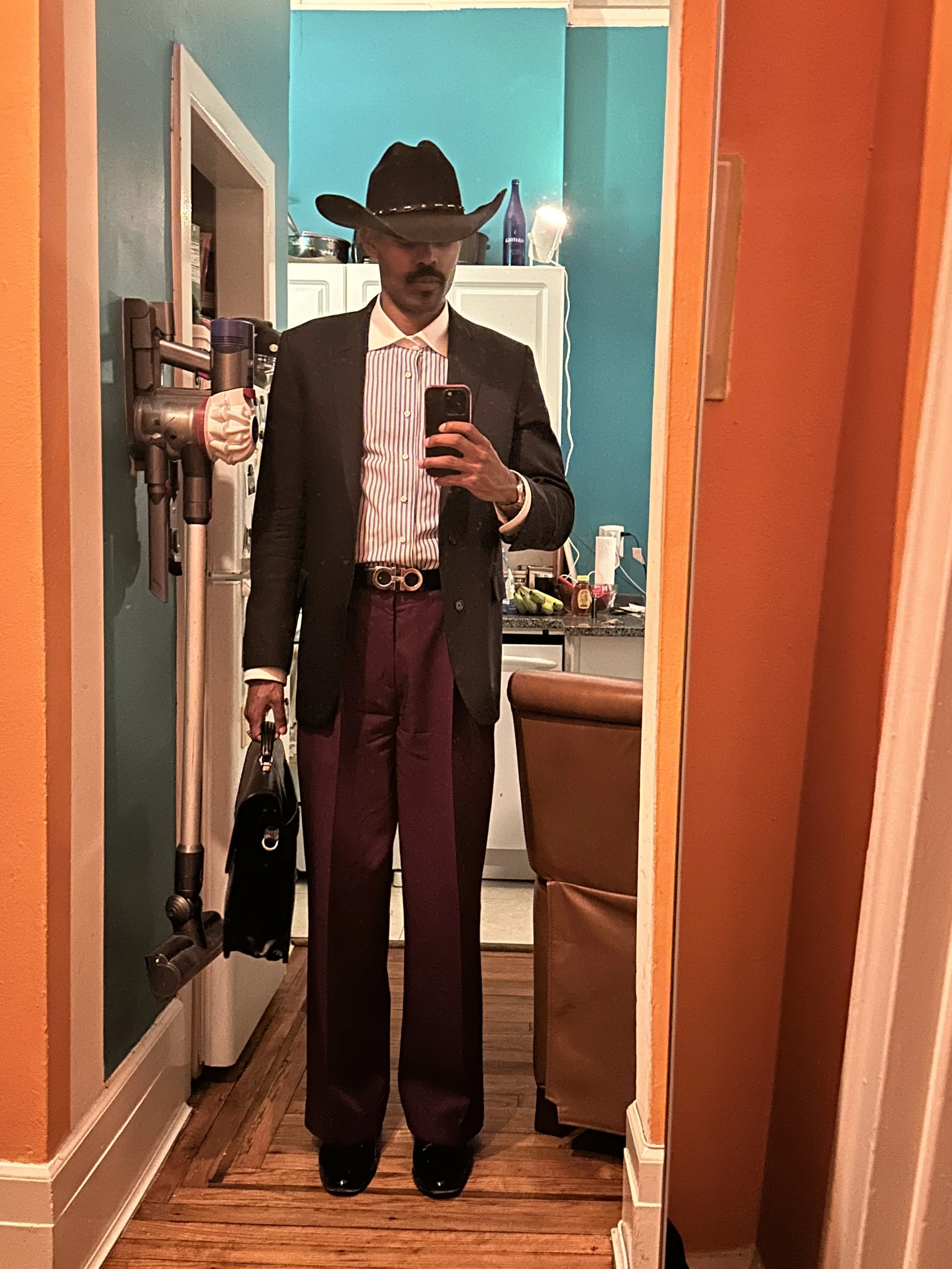 Man dressed in a suit with a cowboy hat taking a mirror selfie in a colorful kitchen, holding a black bag and wearing black shoes.