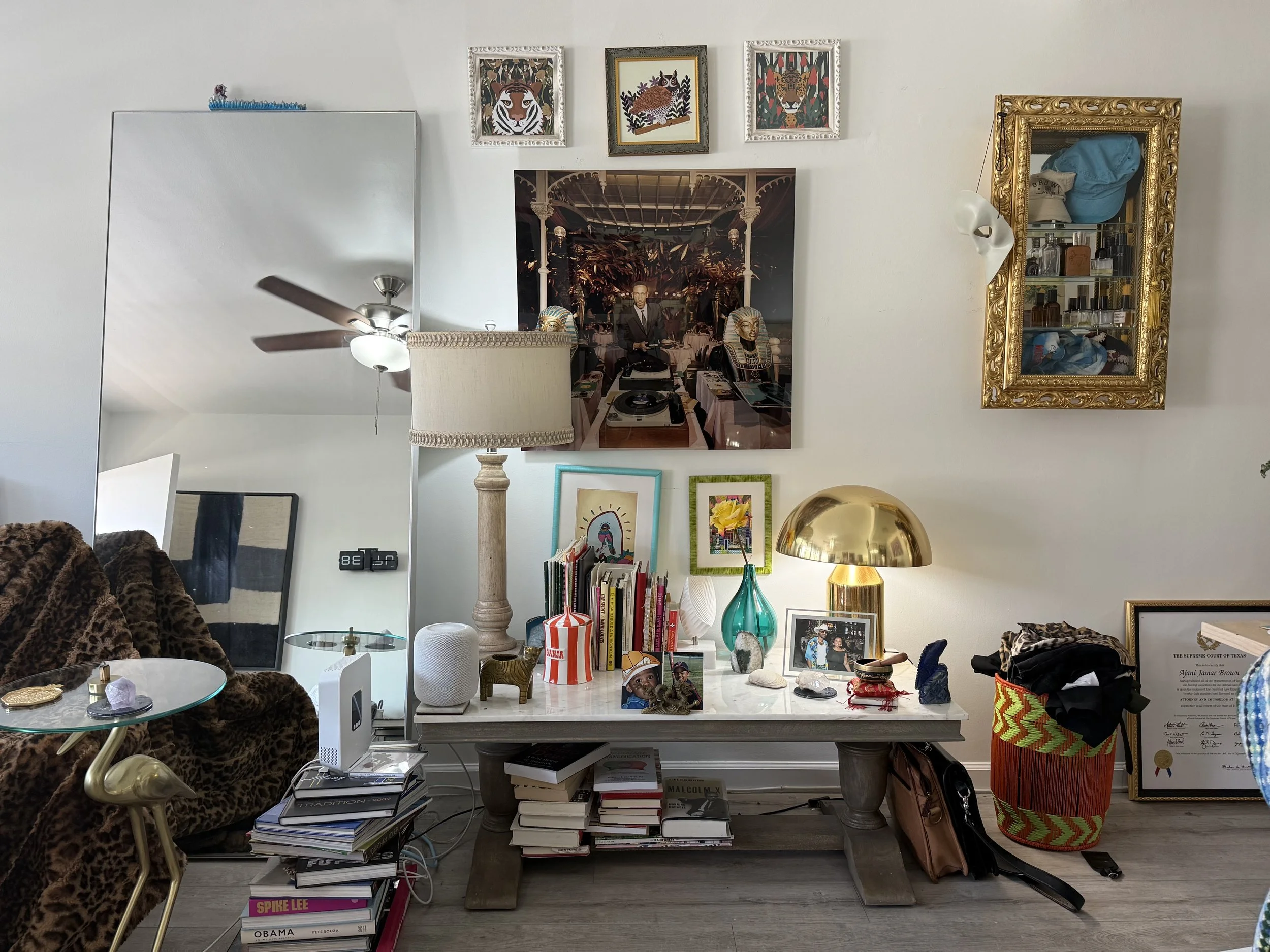 Living room wall with various art pieces, a large mirror, a gold-framed mirror, a table with books, a lamp, vases, and decorative items, and a cluttered basket and framed certificate on the floor.