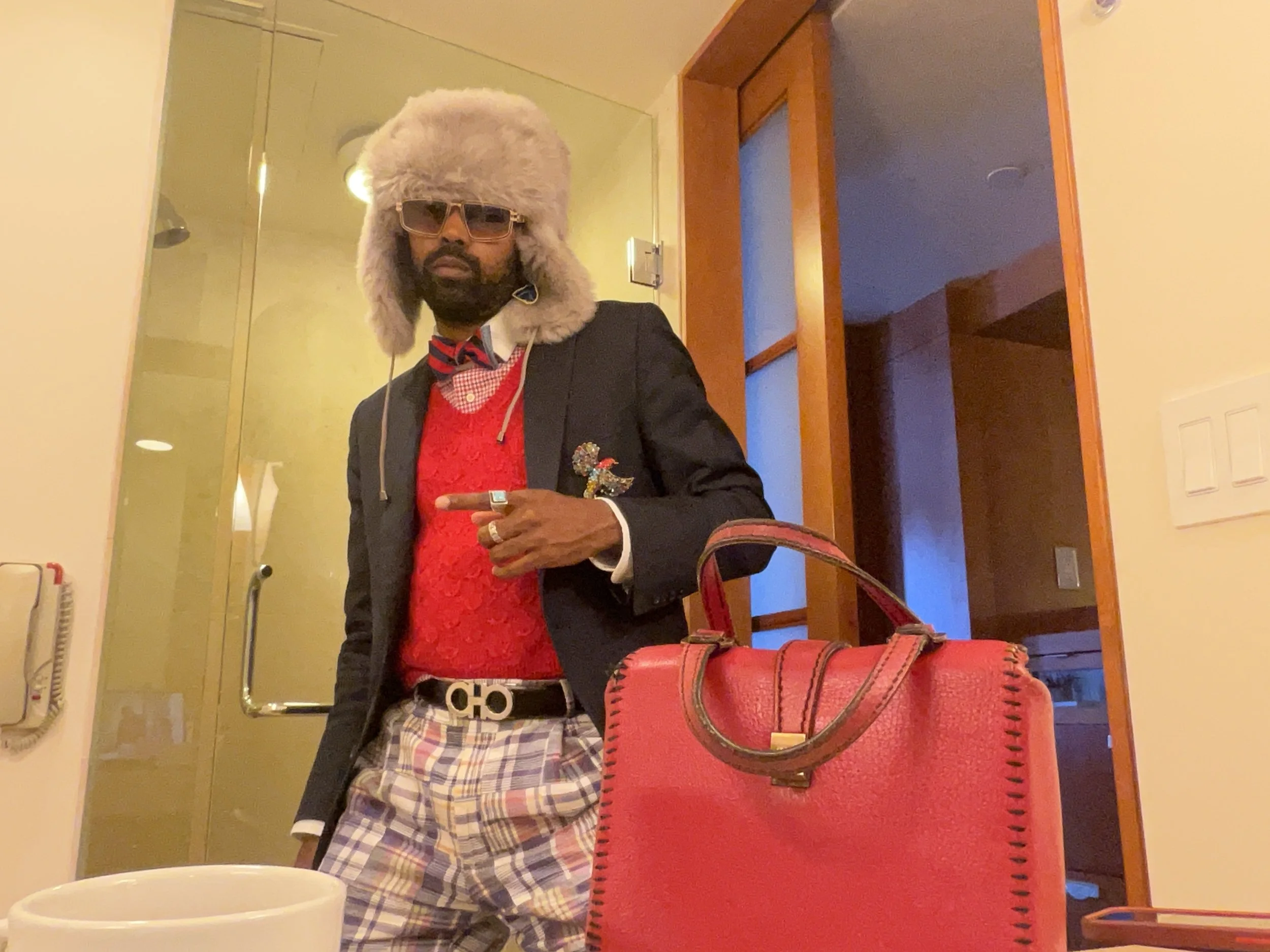 A man stands indoors wearing a large furry gray hat with ear flaps, sunglasses, a black blazer with a decorative pin, a bright red sweater, a plaid shirt, and plaid pants. He is pointing with his right hand at the camera. There is a red handbag on th