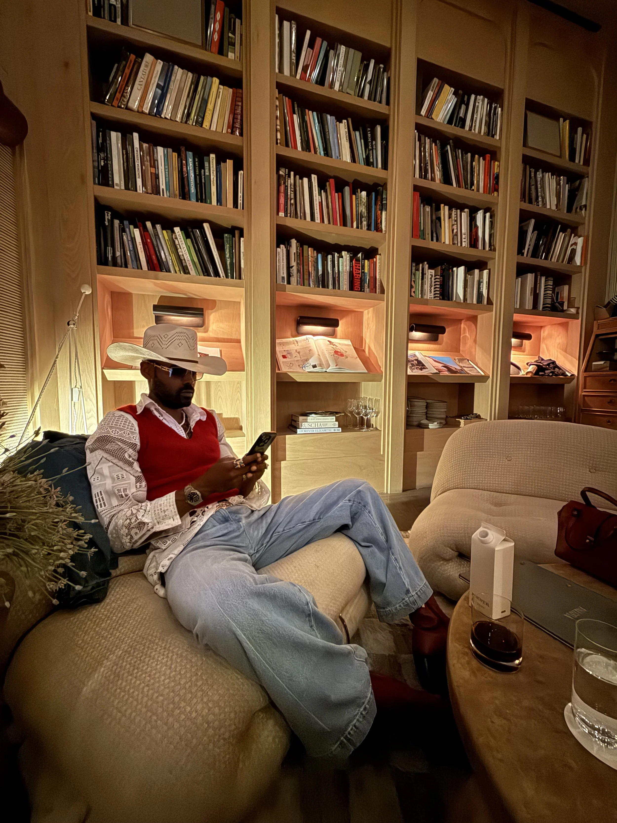 A man wearing a cowboy hat, red vest, white lace shirt, and jeans is sitting on a beige couch, looking at his phone. Behind him is a large wooden bookshelf filled with books, some open and displayed. On a wooden table in front of him are a water glas