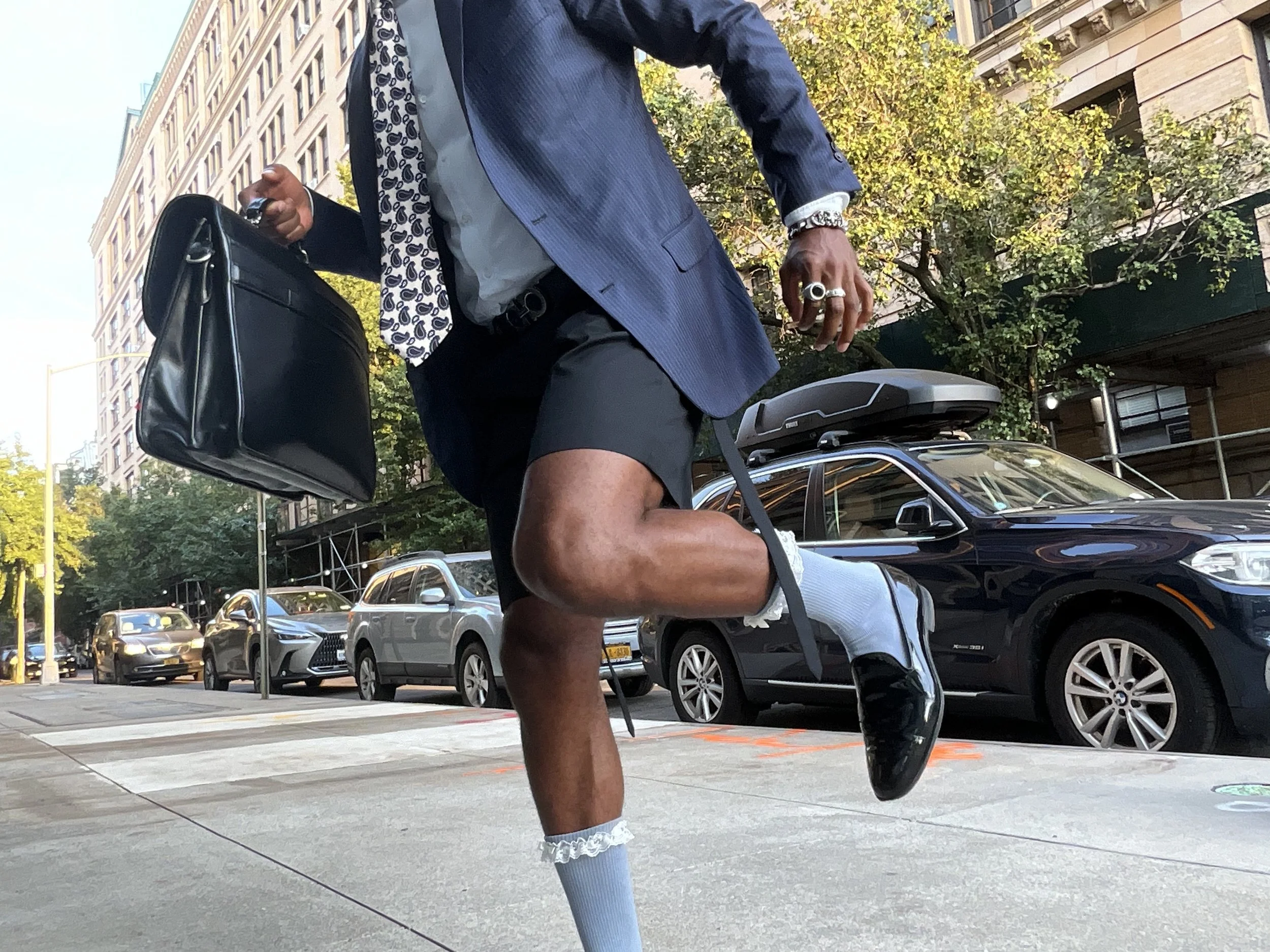 A person walking on a city sidewalk, wearing a blue blazer, dress shirt, shorts, and black shoes with white socks. They are carrying a black briefcase and a black case on their back. The person is also wearing rings, a watch, and a patterned scarf.