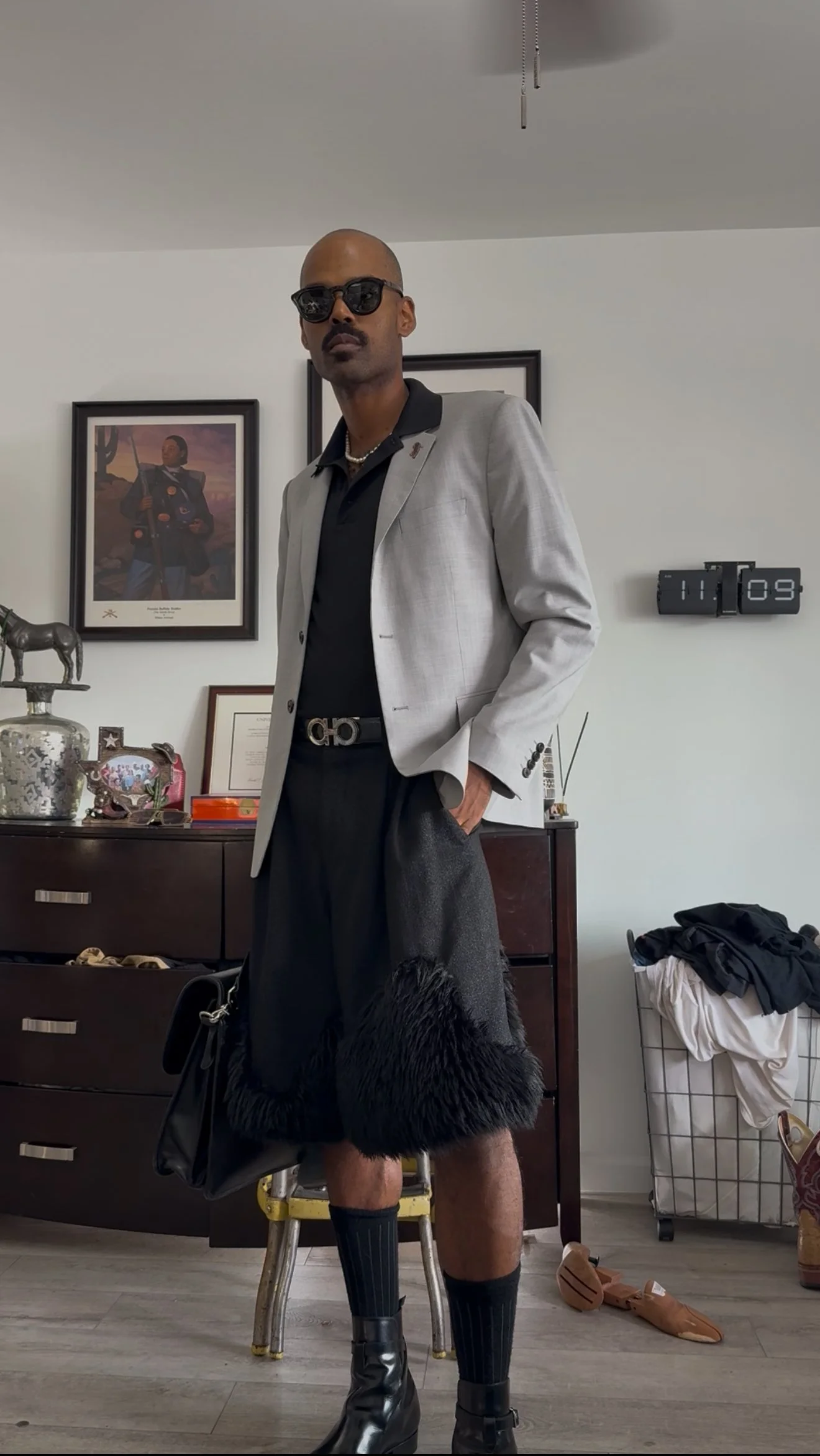 A man in a light gray blazer, black shirt, and black fur-trimmed shorts, wearing sunglasses and standing with his hand in his pocket, in a room with framed artwork, a dresser, and a laundry basket.
