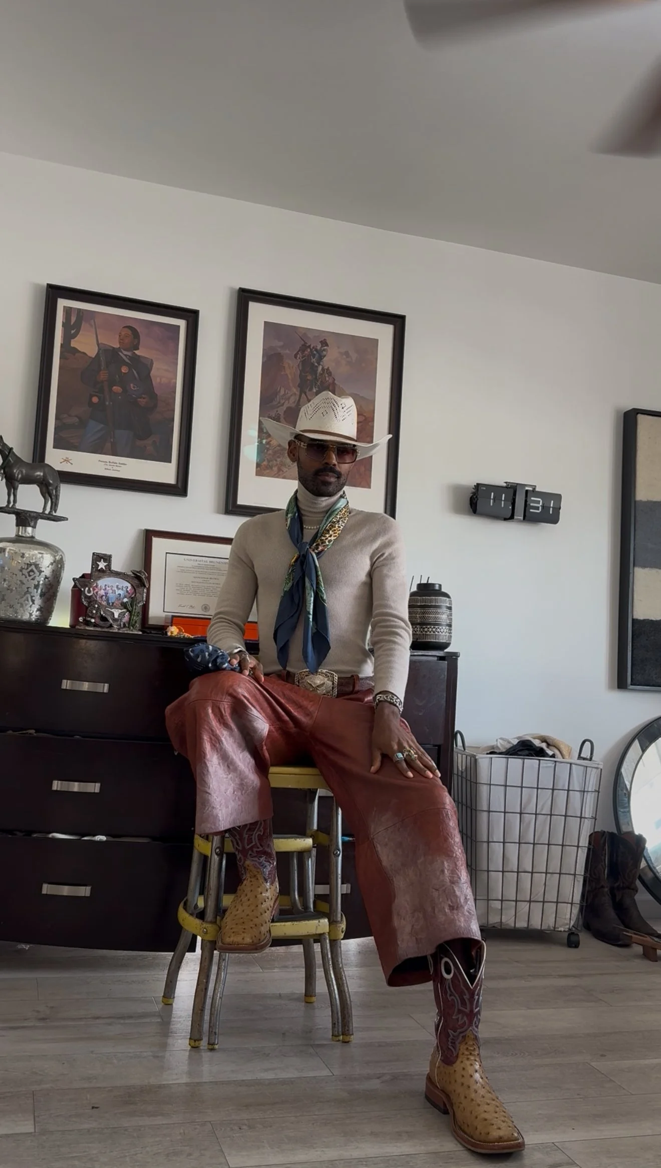 A man dressed in western style clothing with a cowboy hat, sunglasses, a patterned scarf, and cowboy boots, sitting on a chair in a room with western-themed artwork and decor.