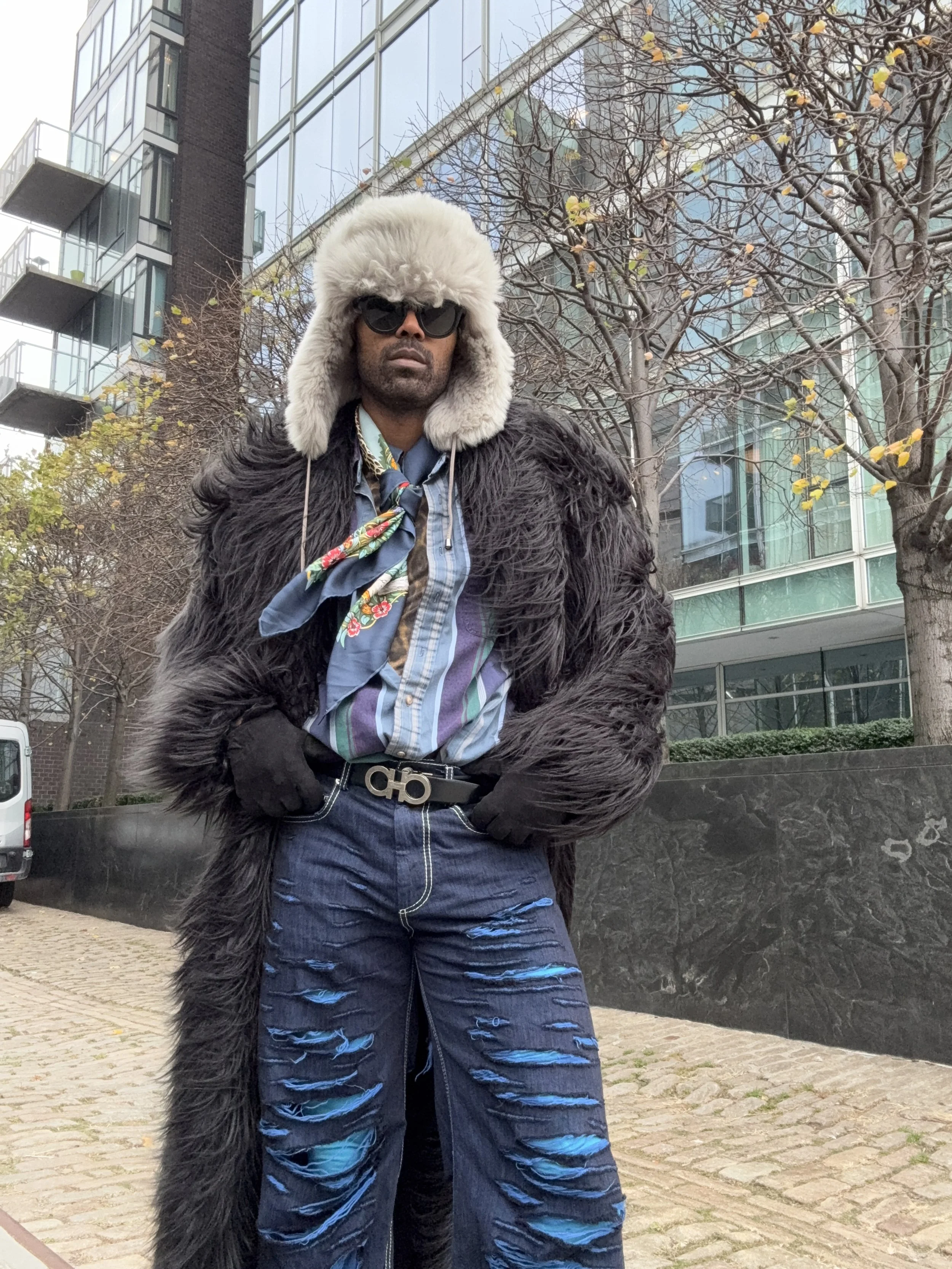 A person standing outdoors in front of a modern glass building and trees with few leaves, dressed in a furry hat and coat, sunglasses, a colorful scarf, and distressed jeans.