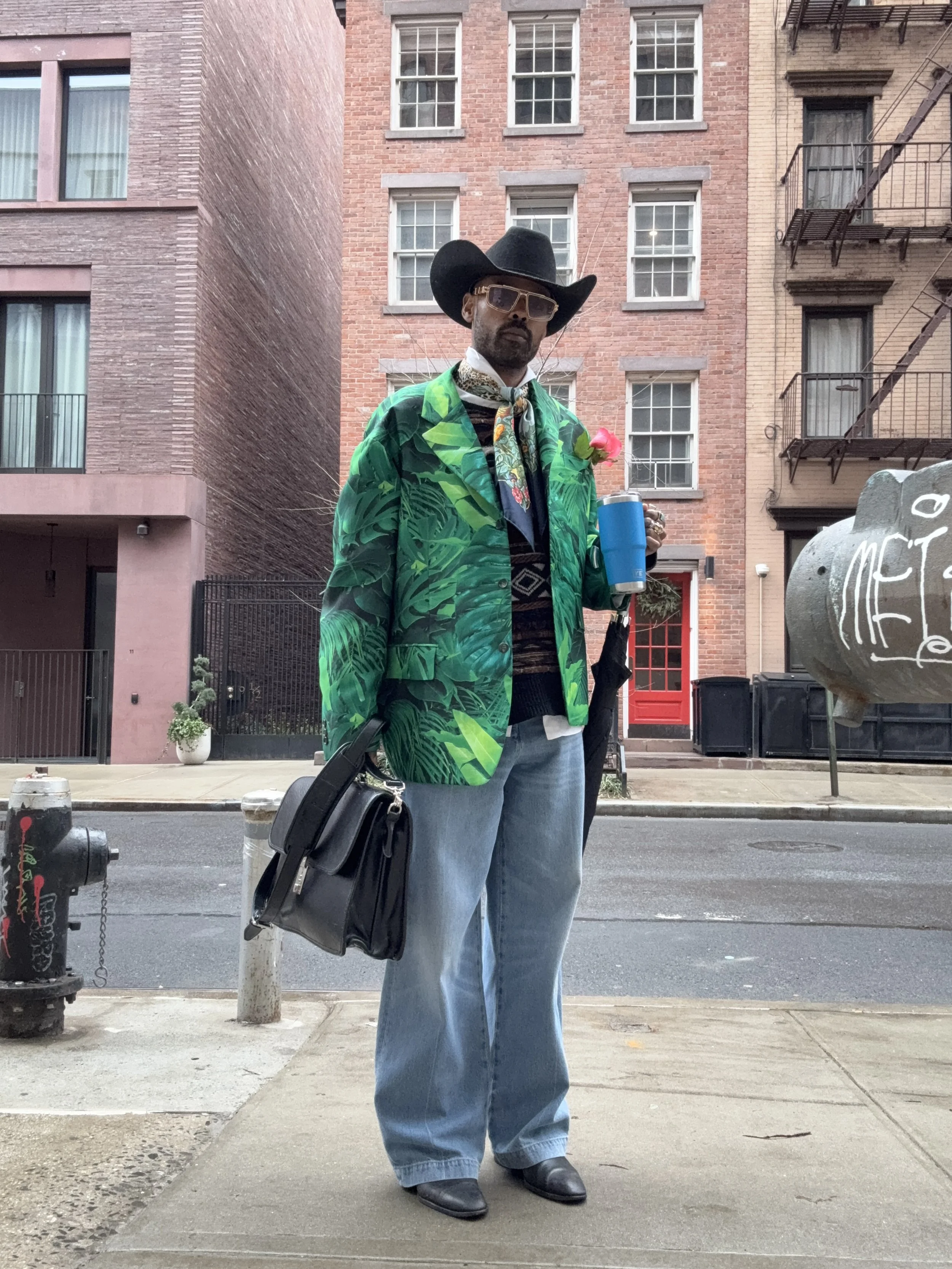 A man stands on a city sidewalk wearing a large black cowboy hat, sunglasses, a colorful tropical print jacket, light blue jeans, and black shoes. He holds a black bag with one hand, a blue cup in the other, and has a pink flower pinned to his jacket