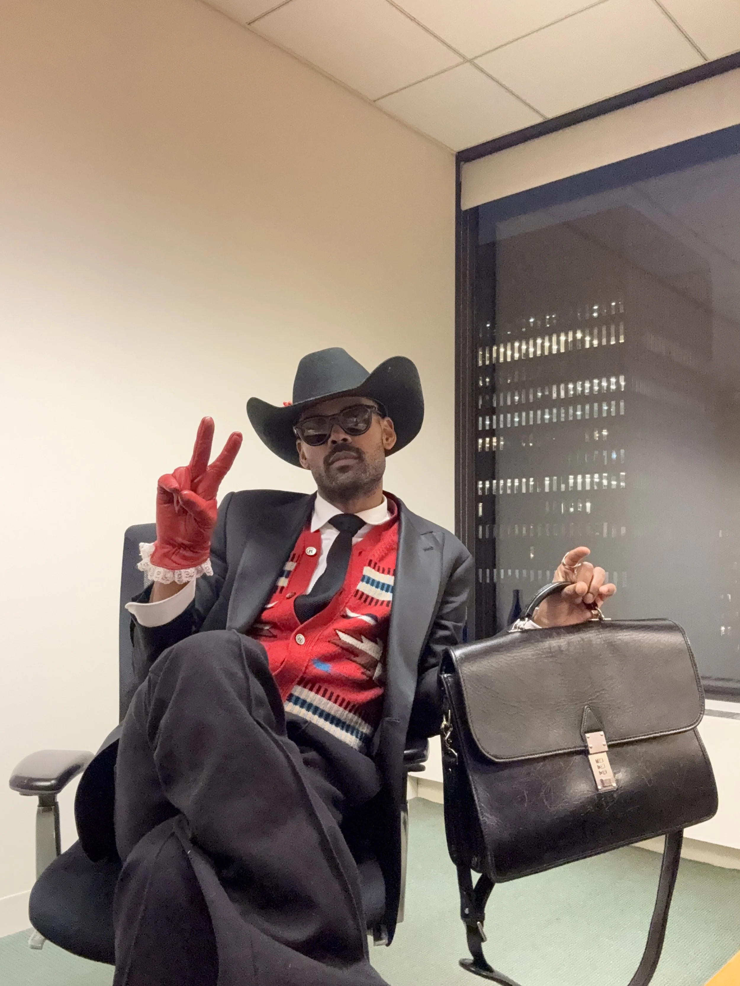 Man dressed in a suit with a cowboy hat and sunglasses, sitting in an office chair, holding a black briefcase, wearing red gloves and making a peace sign with his left hand.