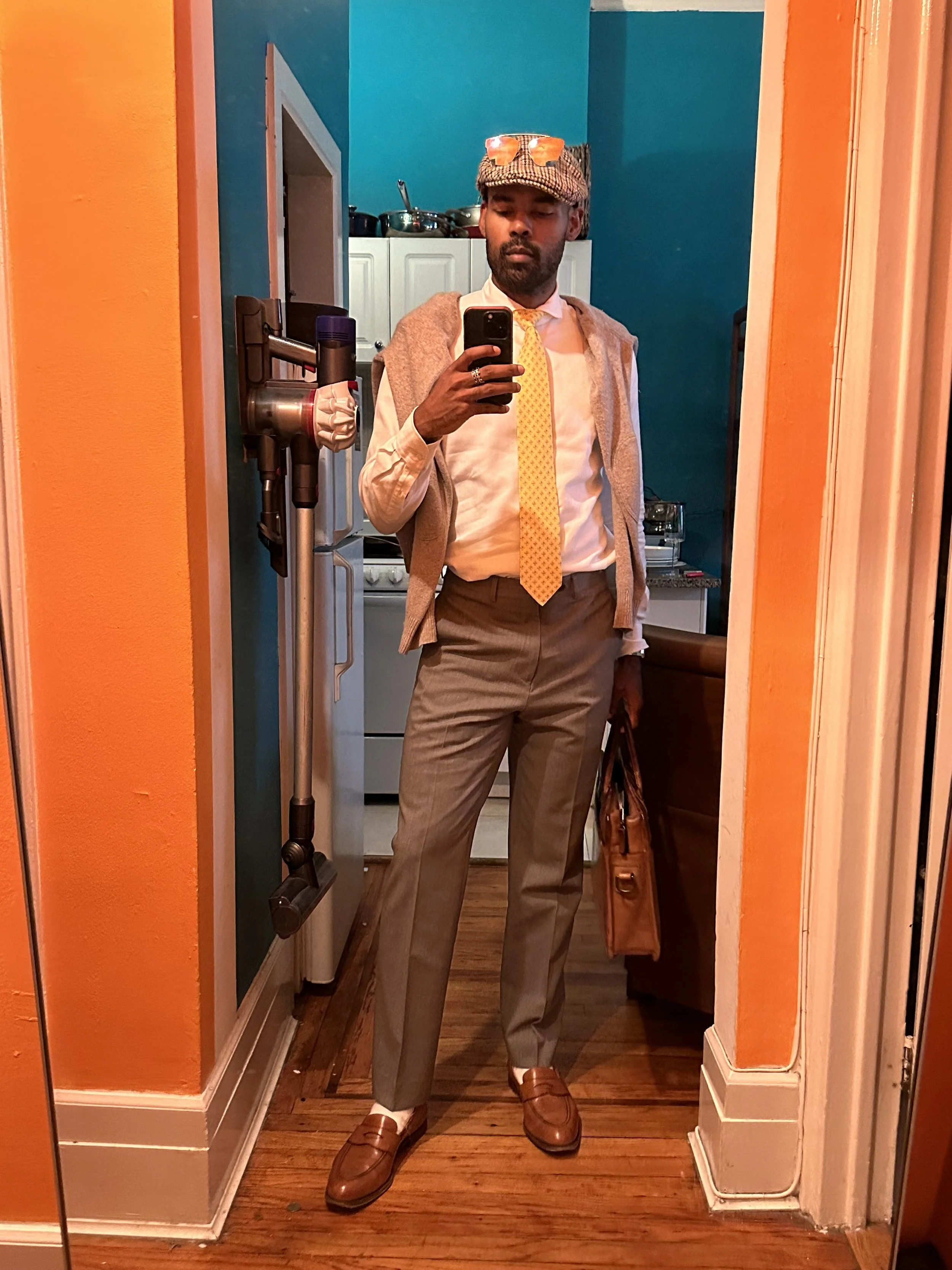 Person taking a mirror selfie in a room with orange and teal walls, holding a brown bag, dressed in a white shirt, yellow tie, tan pants, brown shoes, with a gray sweater draped over shoulders and a patterned hat with orange sunglasses on top.