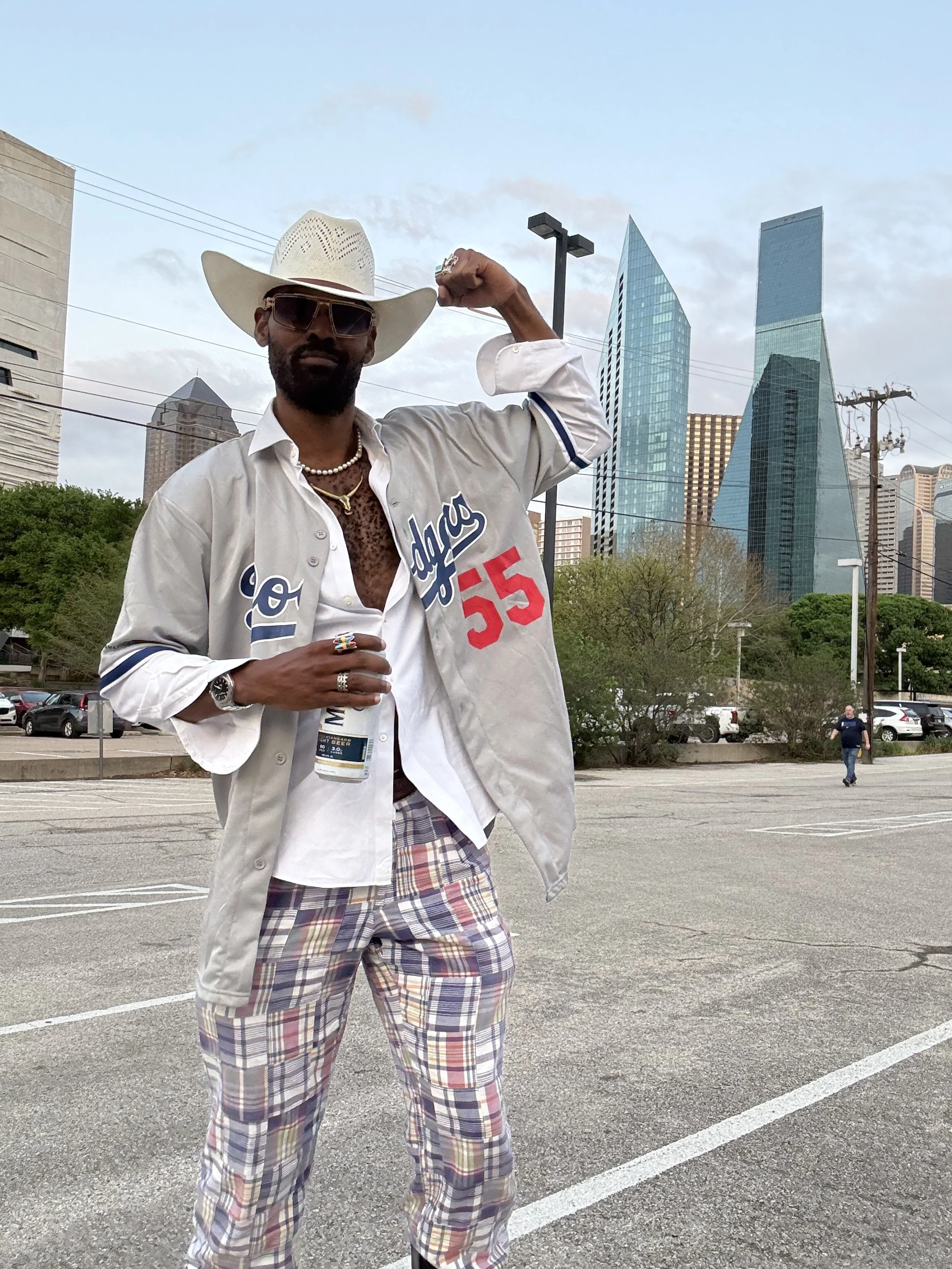 A man dressed in casual and flashy attire standing in a parking lot in front of a cityscape with modern skyscrapers, holding a bottle and flexing his arm.