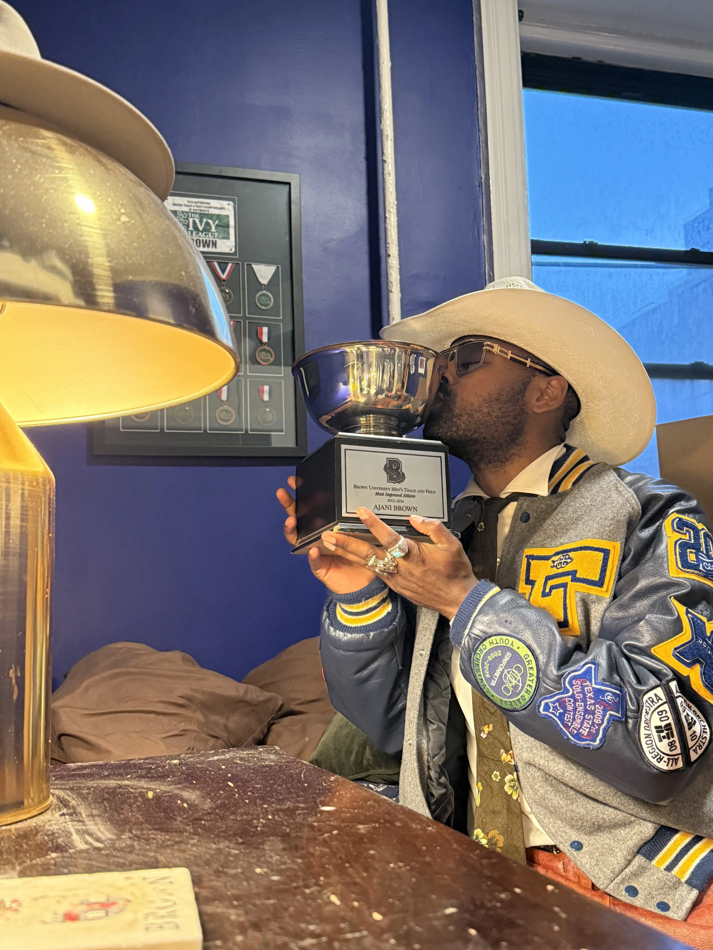 A man wearing a large white hat, sunglasses, and a colorful varsity jacket is kissing a trophy. The trophy has a plaque with the name Ajani Brown. The background includes a blue wall, a framed display of medals, a window, and a lamp.