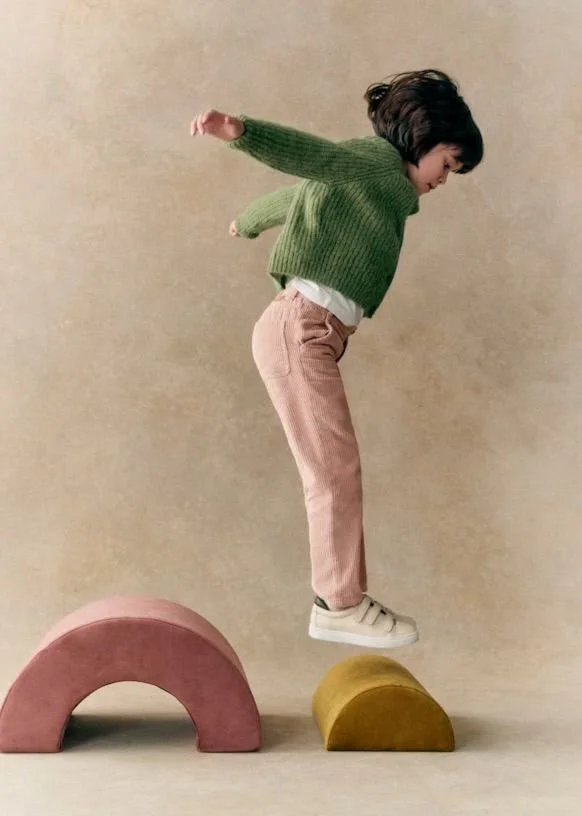 Child balancing on two colorful semi-circular blocks, wearing a green sweater, light pink pants, and white sneakers, with arms extended for balance.