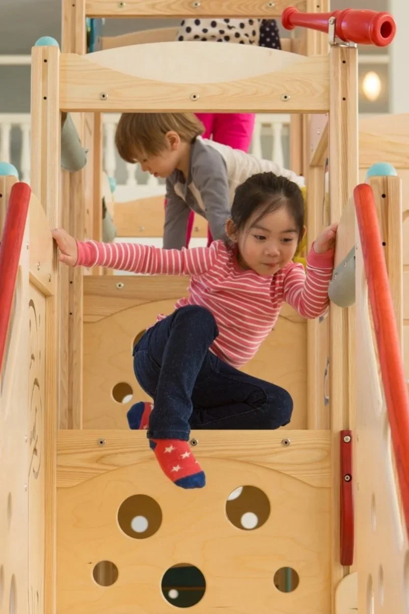 Two children, a girl and a boy, playing on a wooden indoor playground structure with a slide, in a playroom setting.