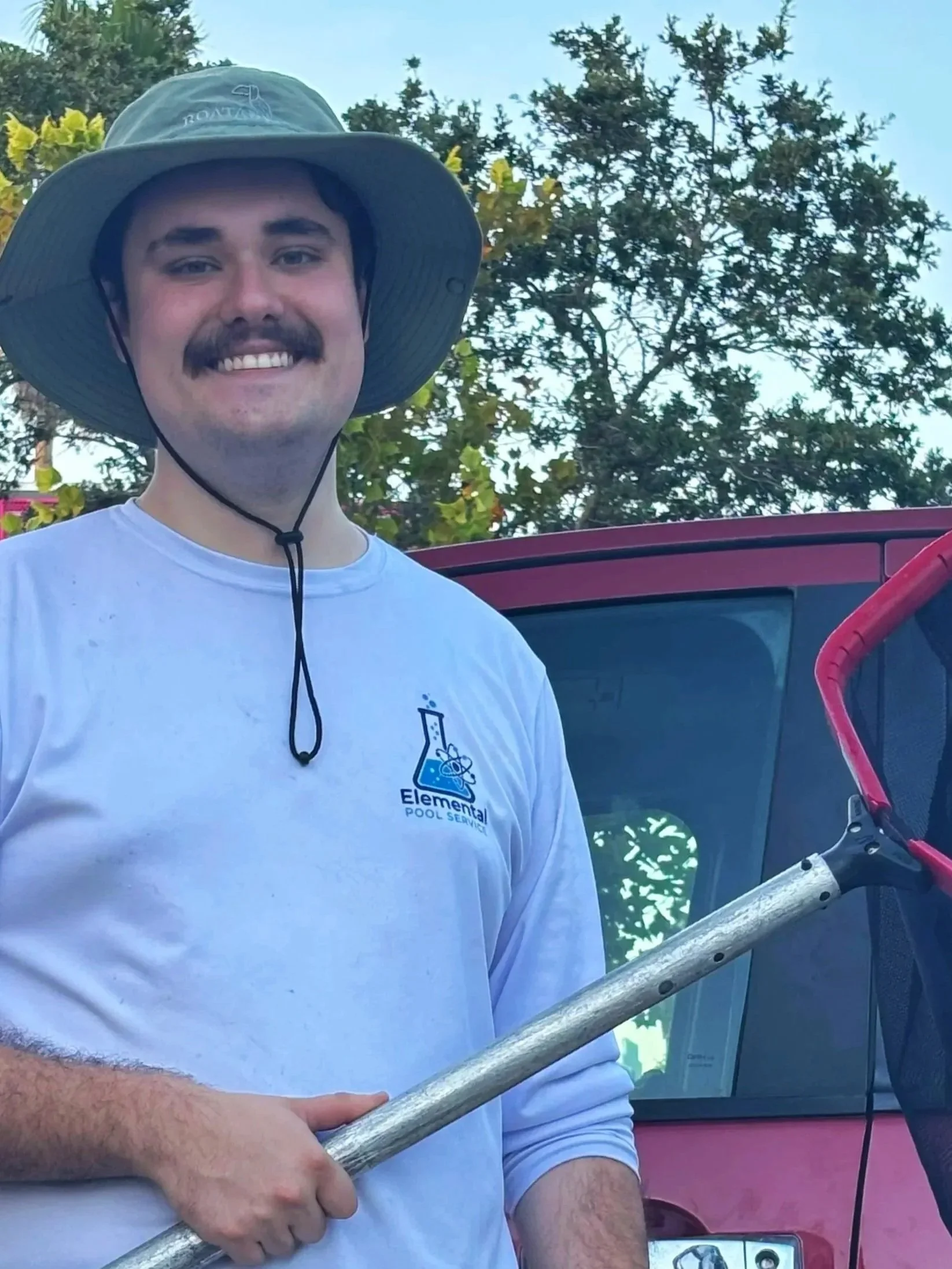 A young man smiling outdoors, wearing a wide-brimmed hat and a white long-sleeve shirt with a pool service logo. He is holding a pool skimmer net, standing near a red vehicle.
