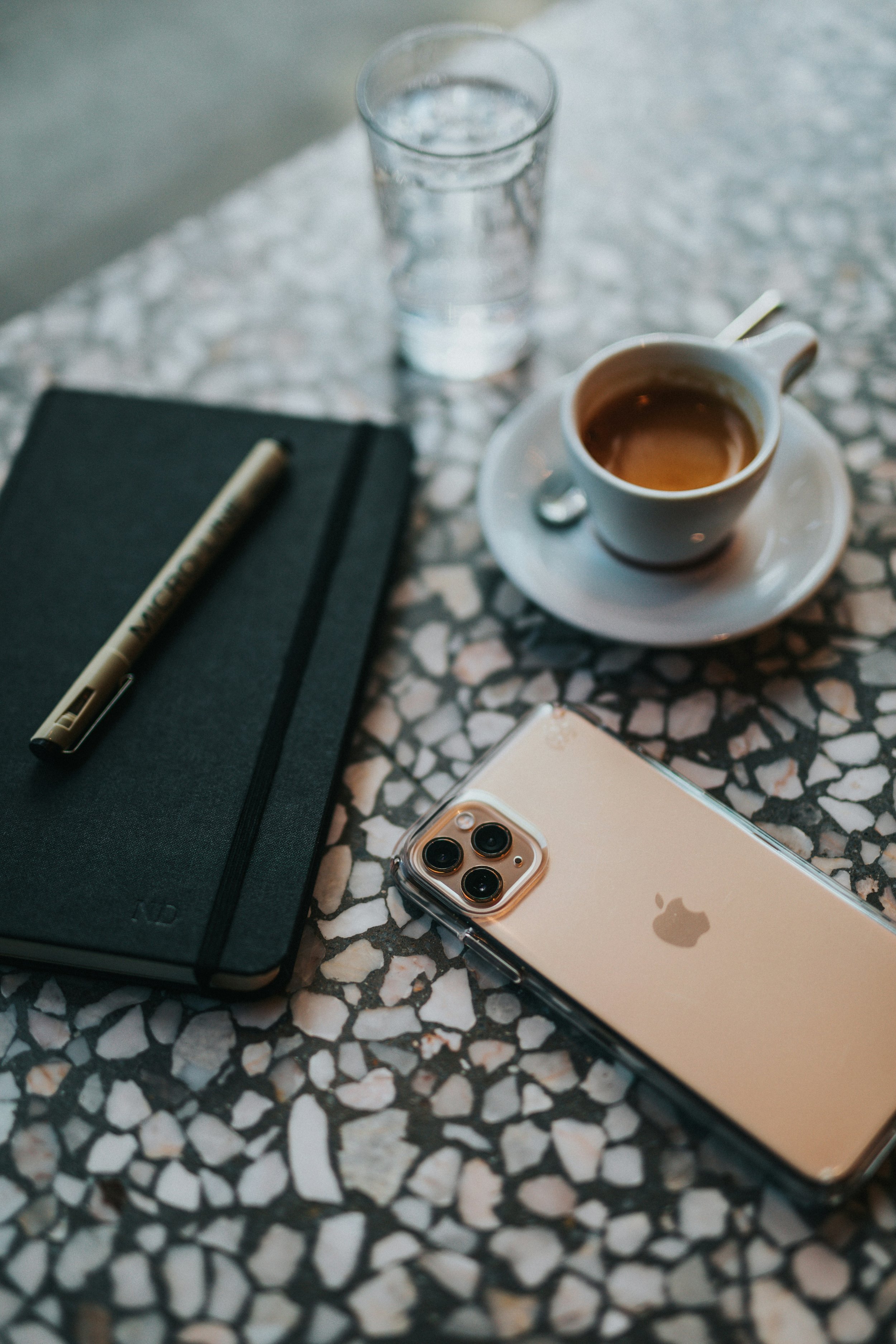 A table with a black notebook, a pen, a glass of water, a white coffee cup with coffee, and a gold iPhone.