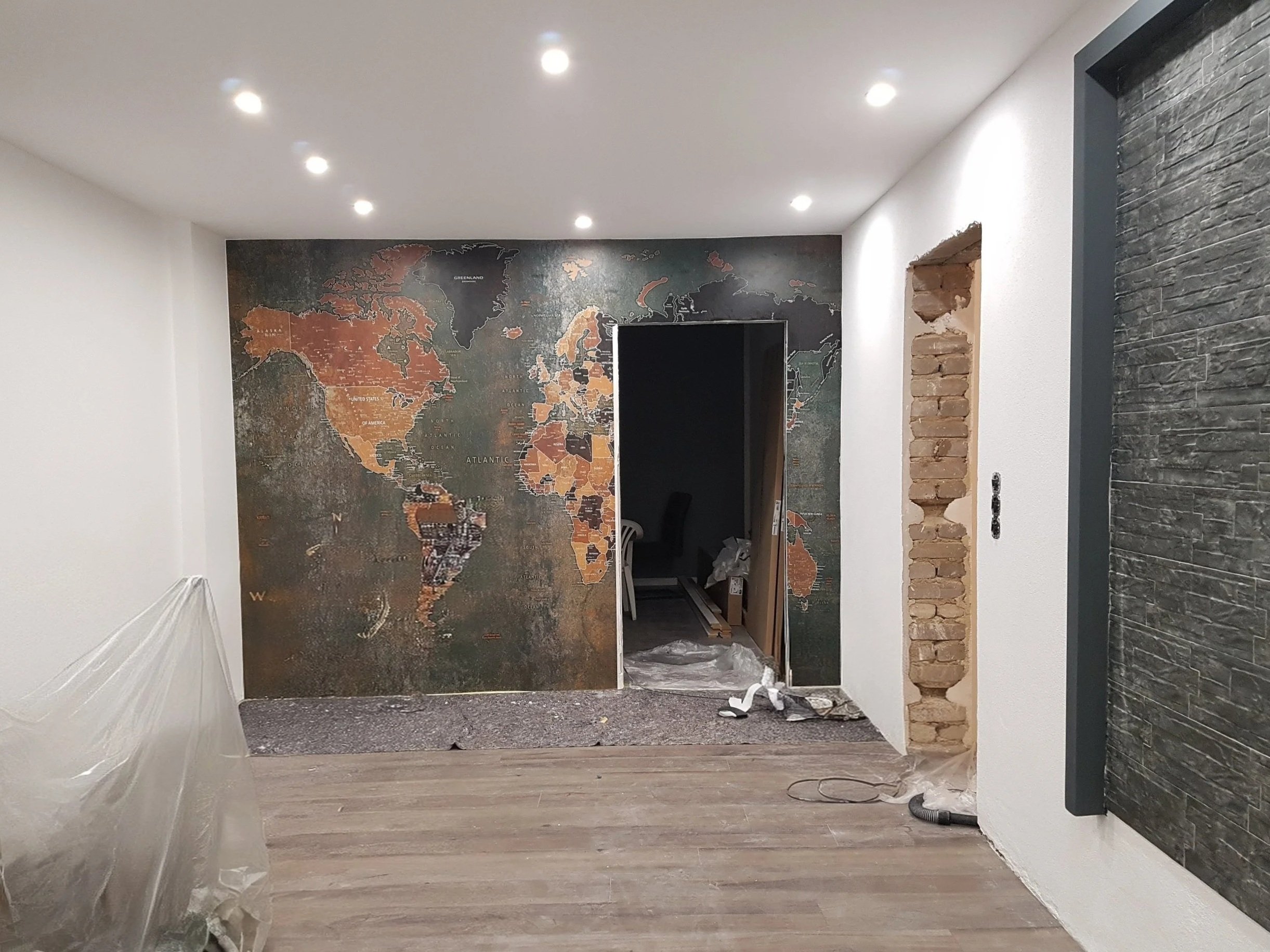 Innenraum mit Weltkartenwand, umrahmter Türöffnung, teilweise sichtbare Ziegelwand, Holzfußboden und Deckenbeleuchtung.