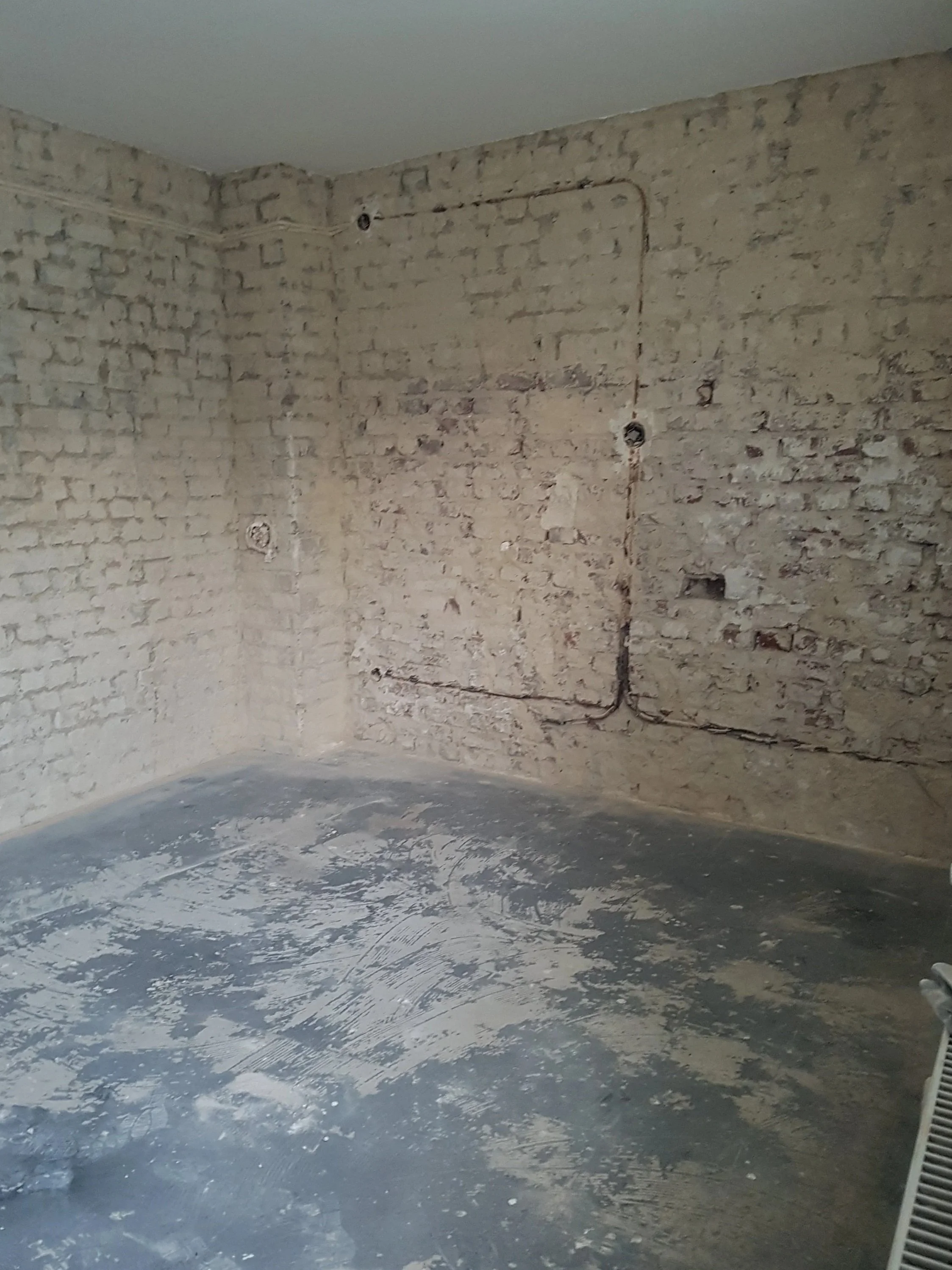Ein unmöbliertes Zimmer mit ungeputzter, bröckelnder Backsteinwand, einem verputzten Betonboden und einem Heizgerät in der rechten unteren Ecke.