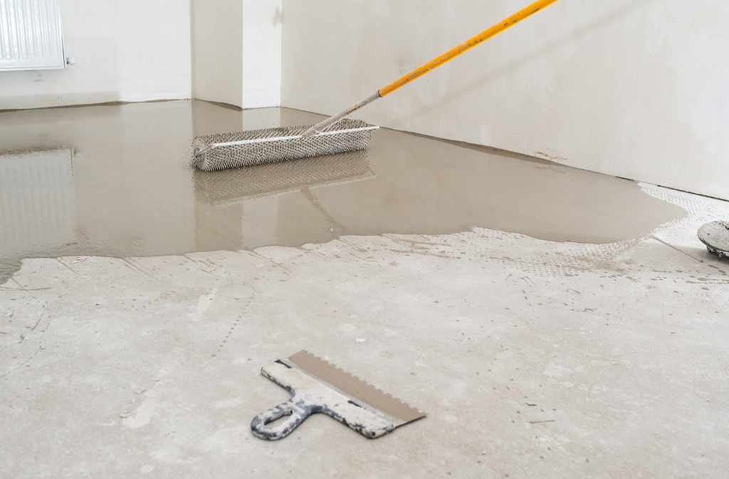 Boden wird mit Beton gegossen, ein Reibebrett und eine Eisenstange sind im Raum zu sehen.