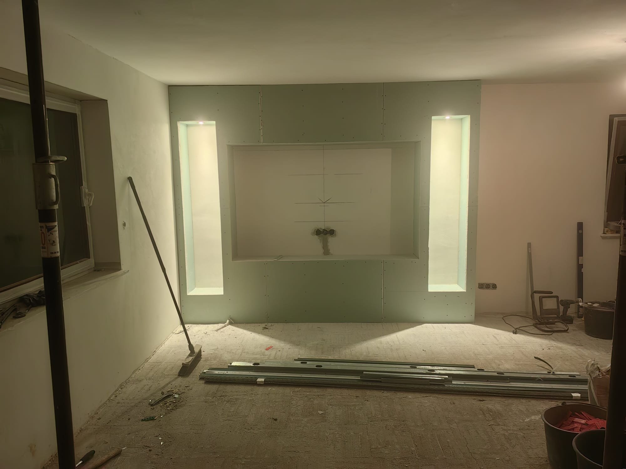 Innenraum eines Raumes, der renoviert wird, mit Baumaterialien und Werkzeugen, eine Wand mit zwei hohen, schmalen Fenstern und eine vorbereitete Aussparung für eine Küche.