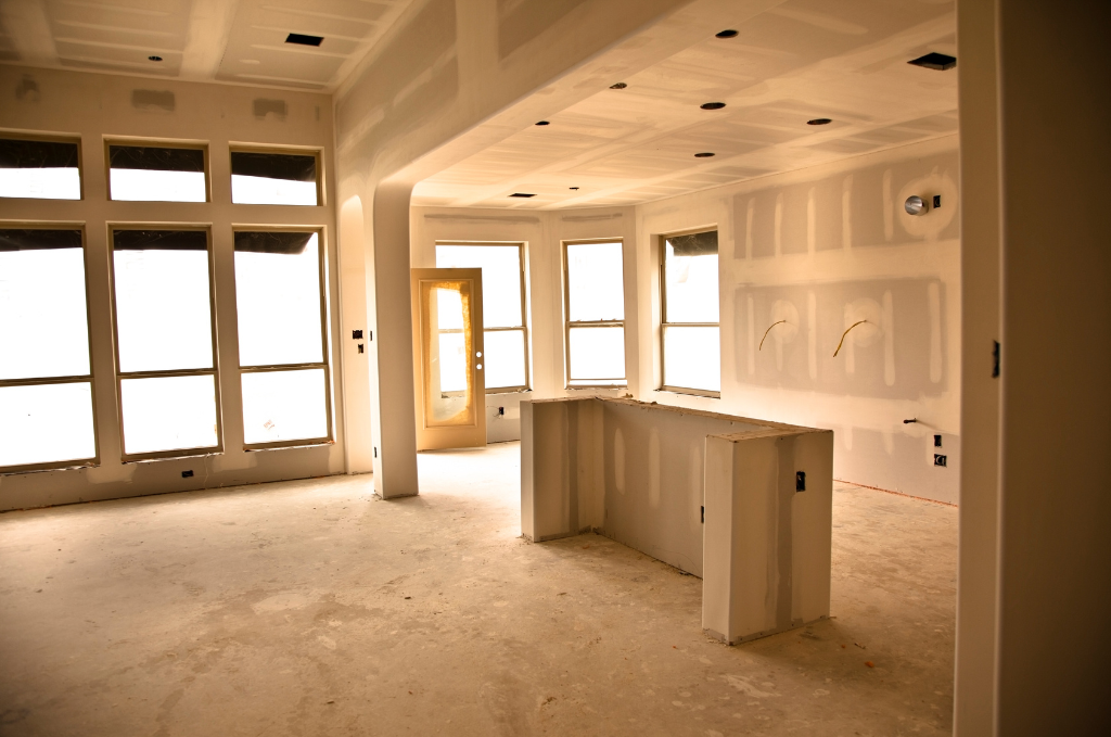 Innenraum eines unfertigen Hauses mit großen Fenstern, unvollendeter Wandgestaltung und einer Kücheninsel aus Trockenbau.