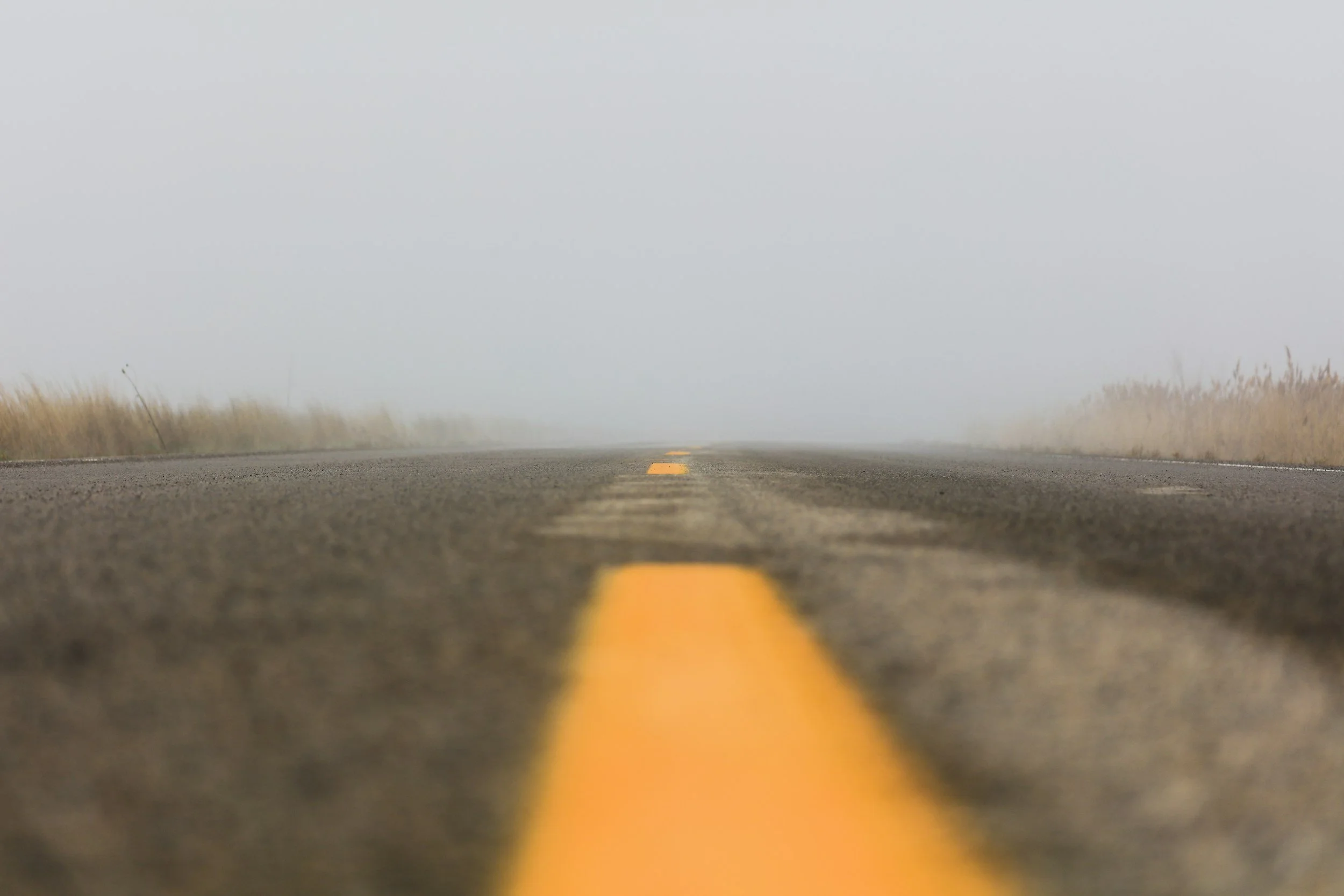A straight, empty road with yellow lines in a foggy landscape, minimal visibility.