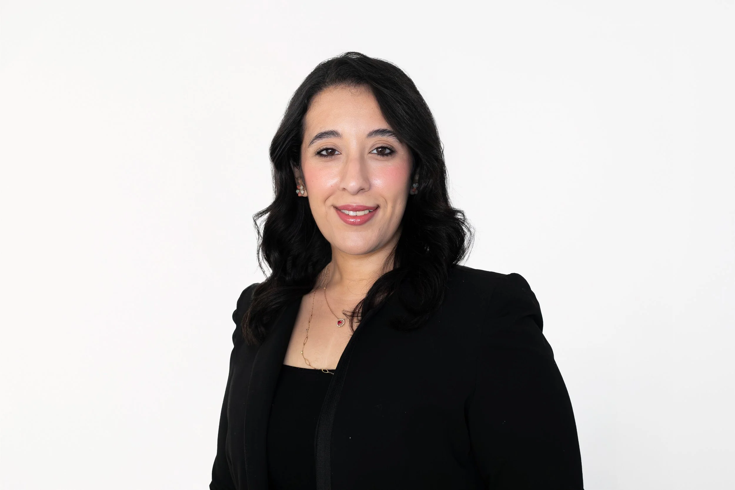 Portrait of a woman with dark hair, wearing a black blazer, jewelry, and a subtle smile, against a white background.