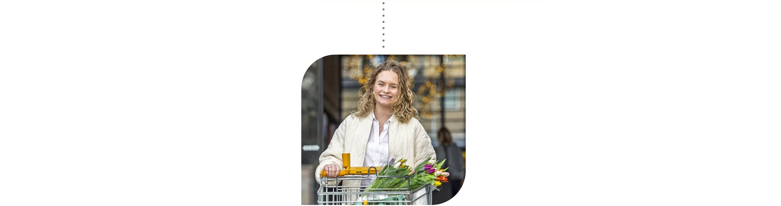 Vrolijke vrouw met curly haar die bloemen in een supermarktkarretje heeft, met een achtergrond van een moderne winkel en mensen.