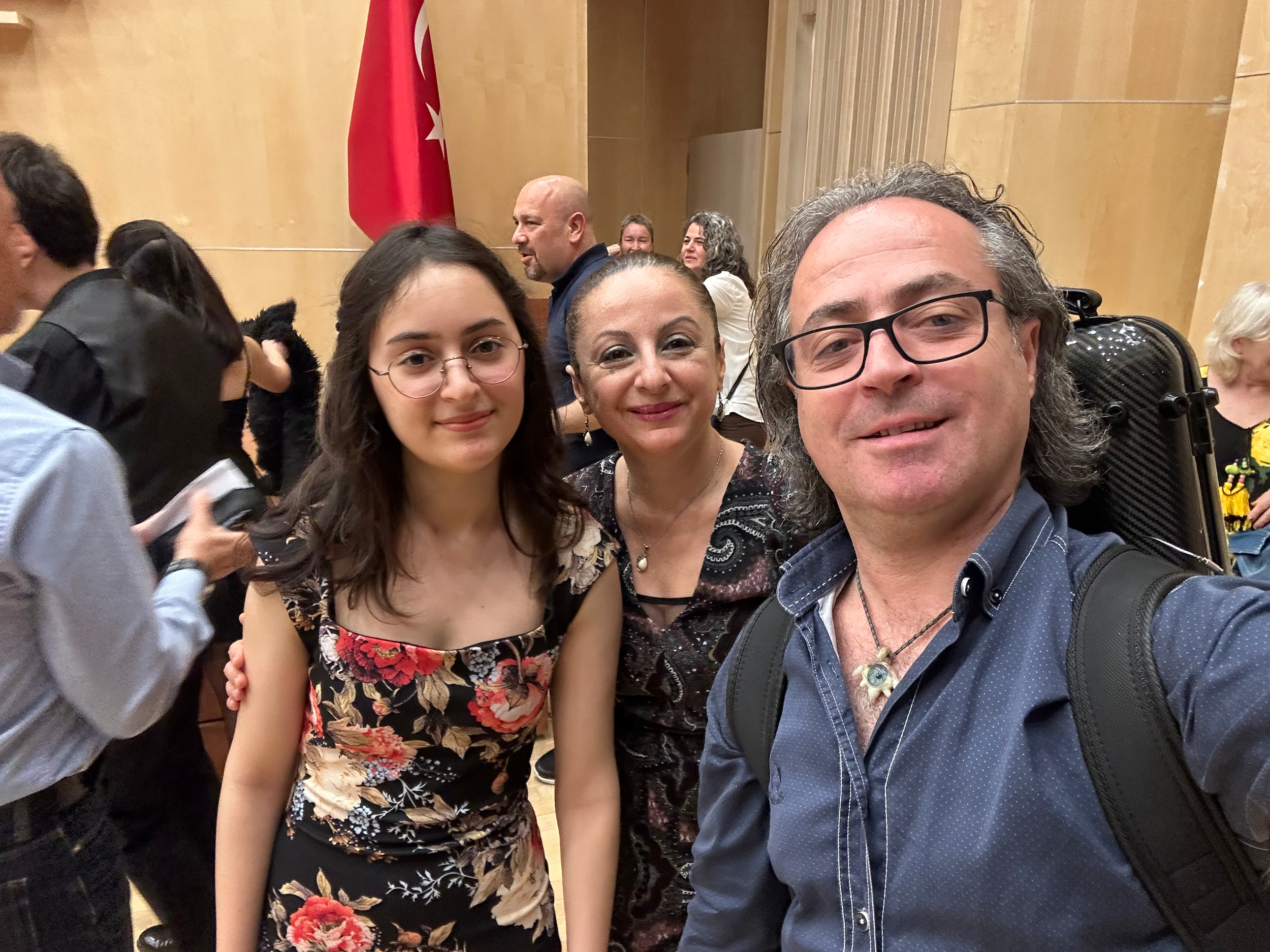 Drei Menschen sitzen nebeneinander und schauen in die Kamera. Zwei Frauen und ein Mann, alle tragen Brillen. Der Mann hat graues Haar und trägt ein blaues Hemd. Die Frauen haben braune Haare, die eine trägt ein schwarzes Kleid mit Blumenmuster, die a