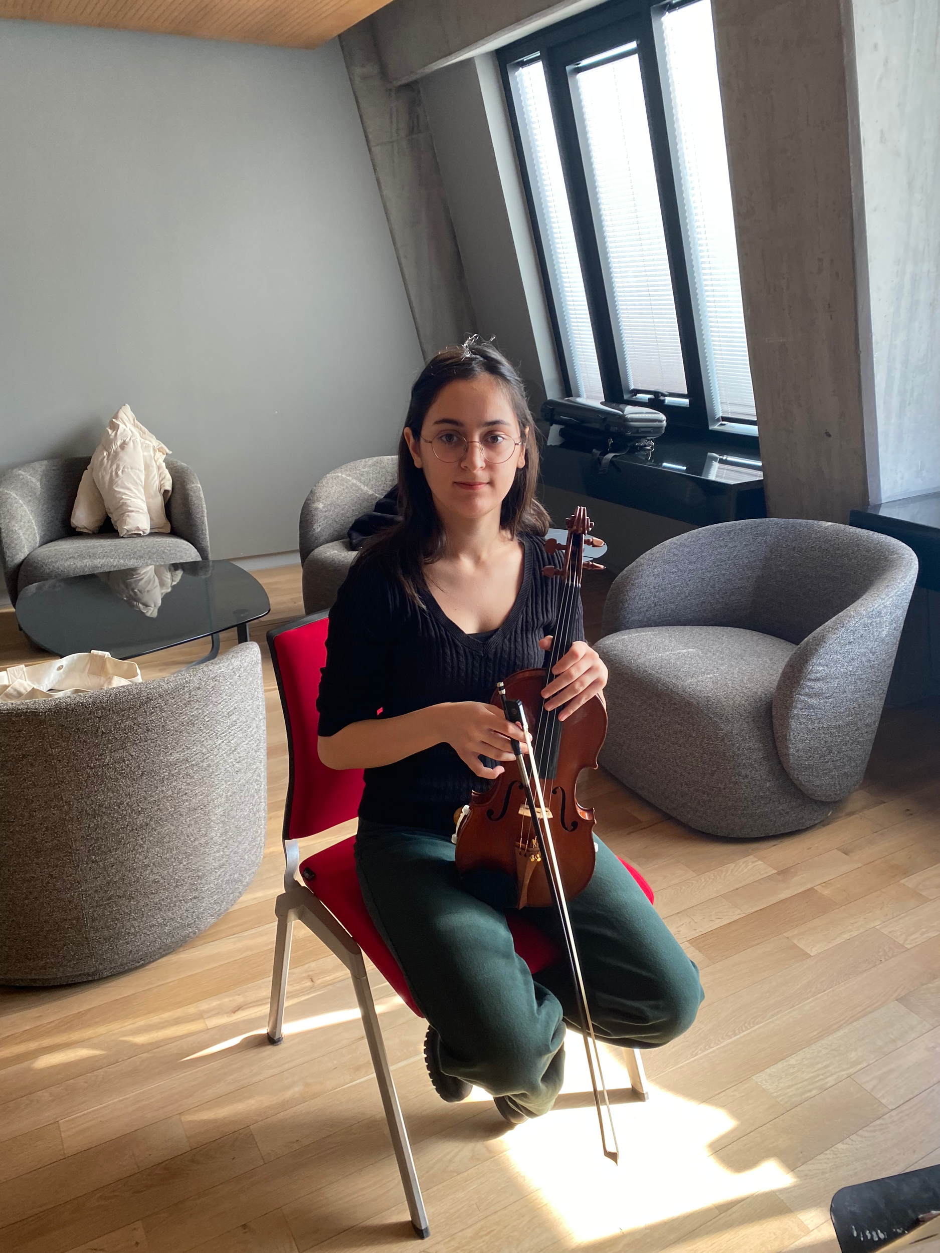Eine junge Frau mit dunklen Haaren, Brille und dunklem Oberteil sitzt auf einem roten Stuhl in einem modernen Raum, hält eine Violine in den Händen, vor einem großen Fenster mit geschlossenen Augenlappen, im Hintergrund graue Sessel und ein Tisch.