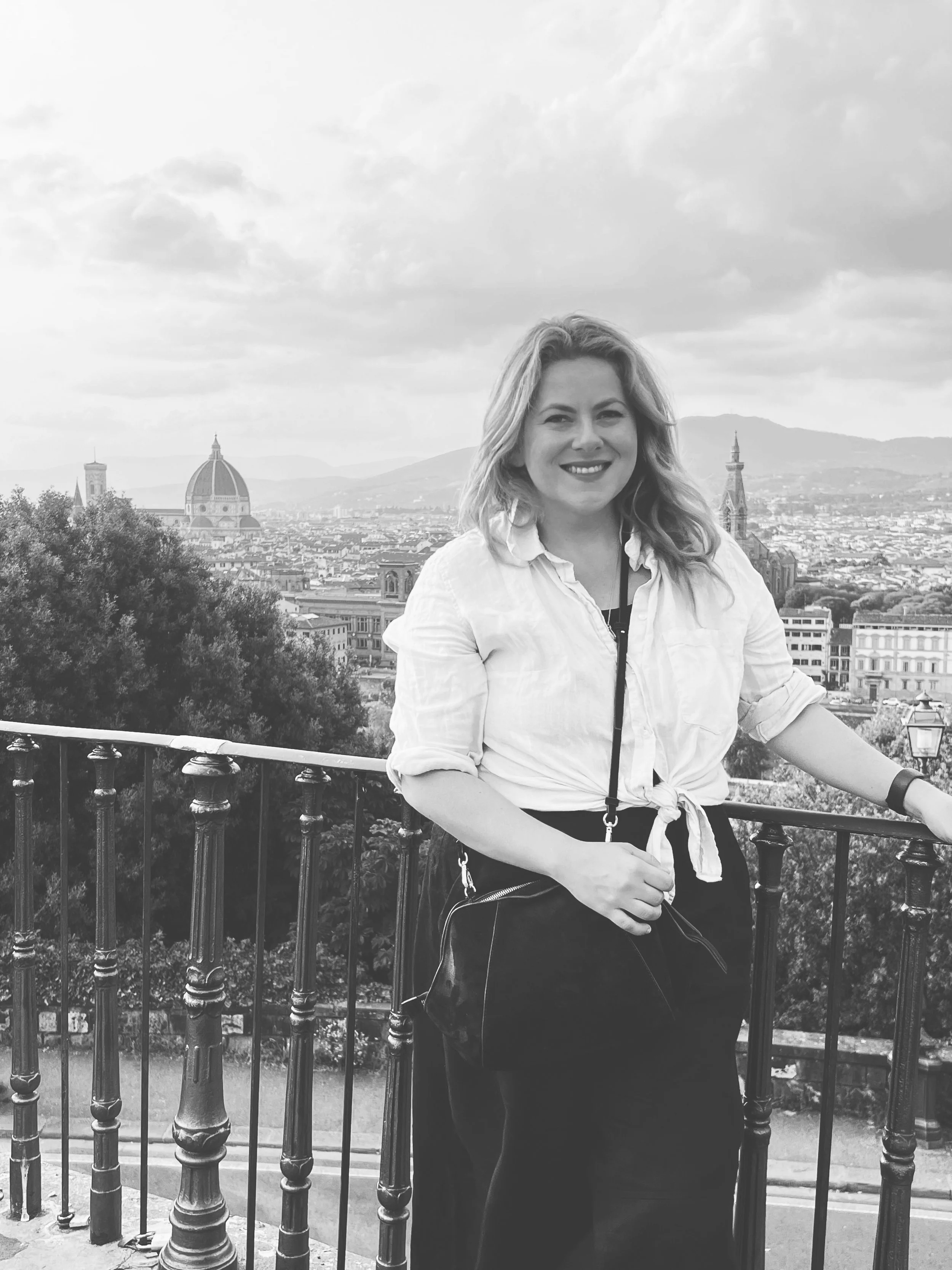 Black and white photo of a smiling woman with shoulder-length hair standing near a railing, with a cityscape including domed and spire buildings behind her, under a cloudy sky.
